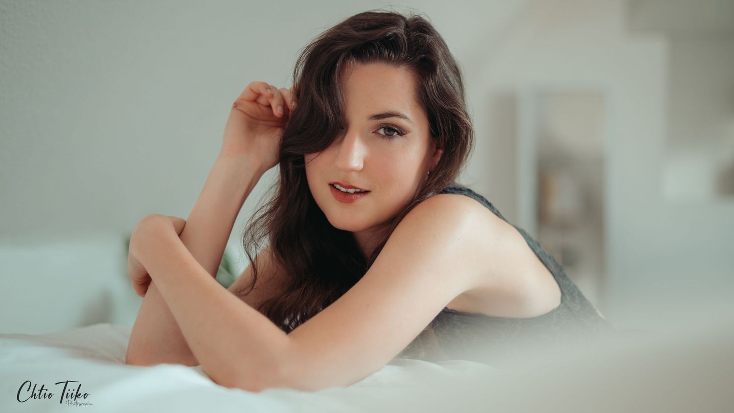 A young brunette woman poses gracefully on a white bed, looking at the camera with a subtle smile.