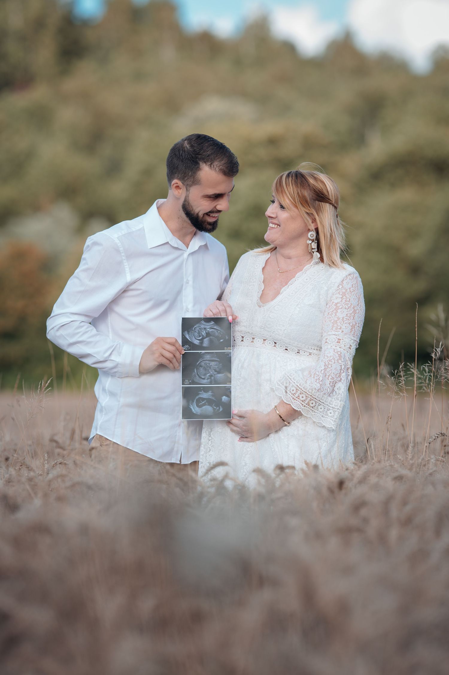 Couple holding ultrasound photos in a wheat field, woman in white lace dress touching baby bump, maternity announcement.