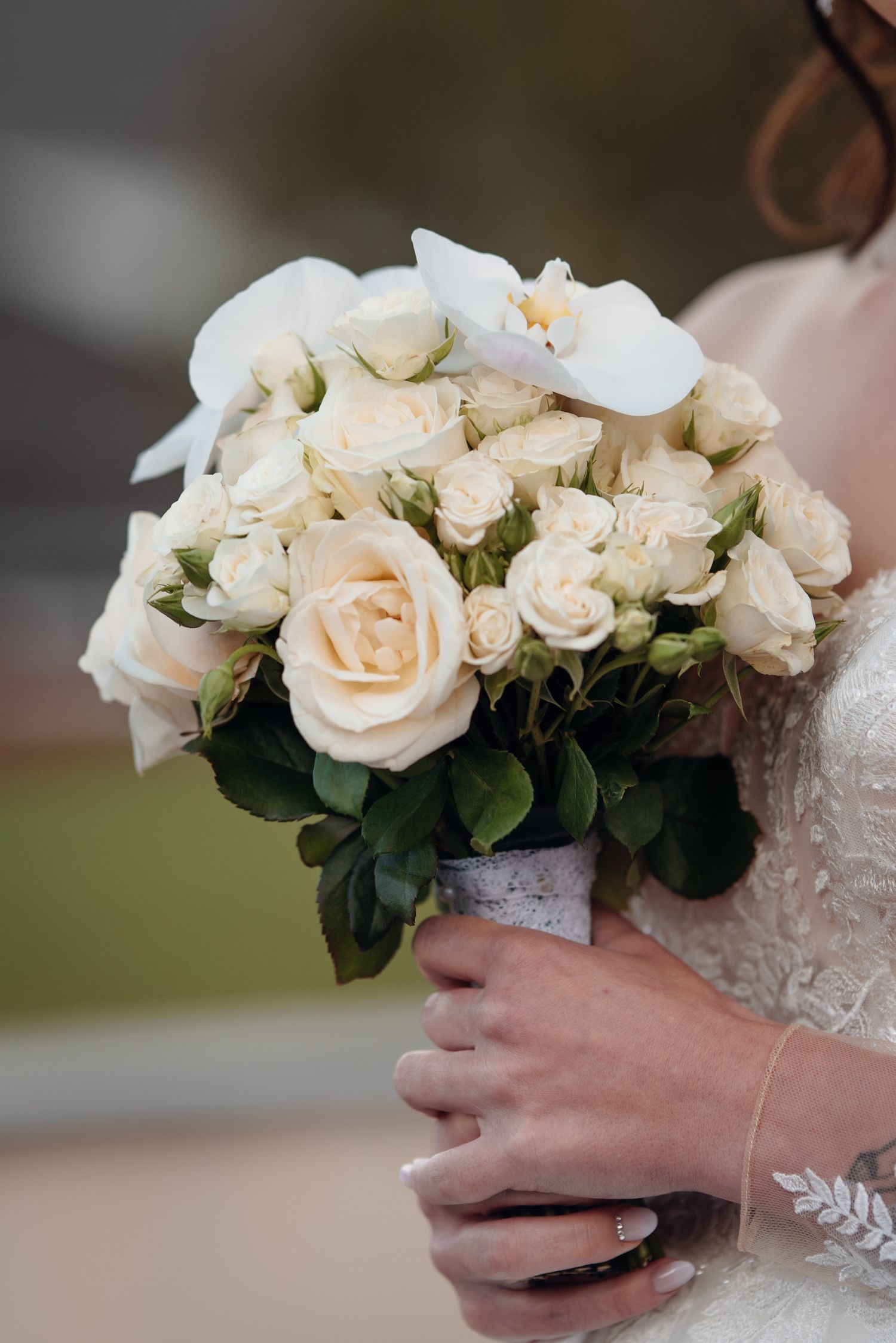 Bride in lace wedding dress holding elegant bouquet of cream roses and white orchids with ring visible.
