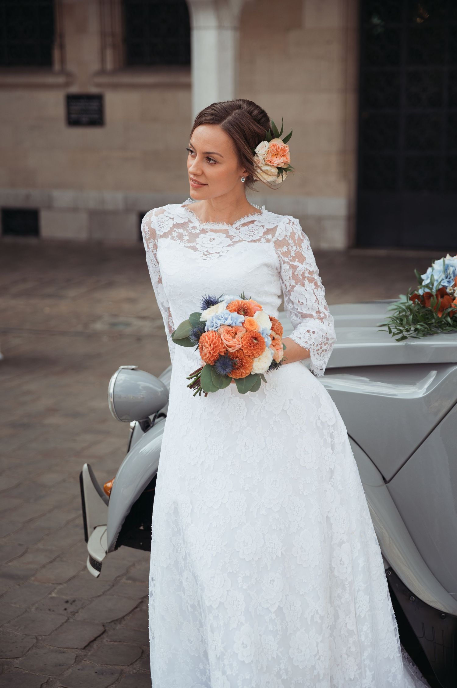 Bride in white lace long-sleeve gown holding orange bouquet, leaning against vintage car outdoors.