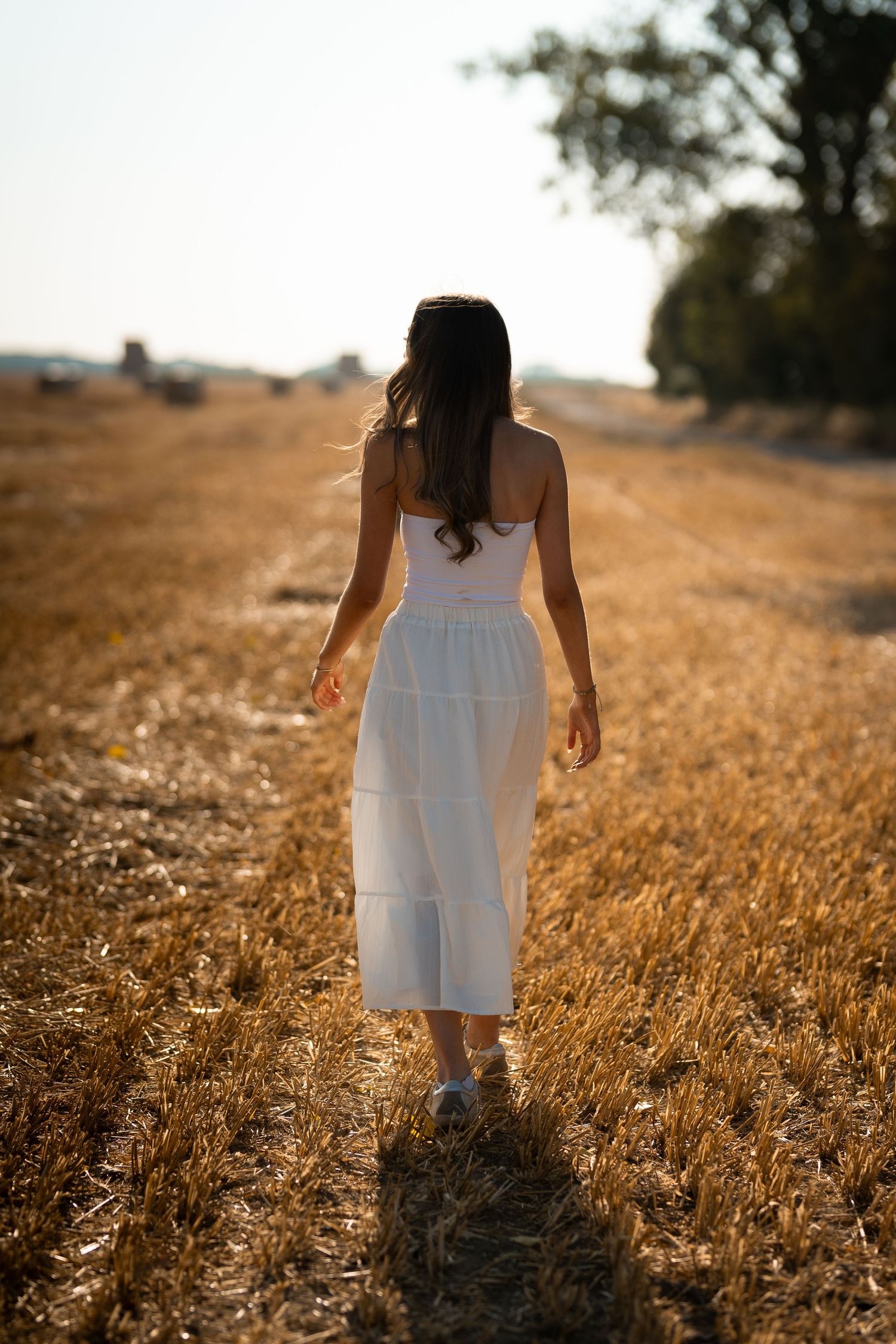 Woman in white dress walking through golden wheat field at sunset, viewed from behind with trees on horizon.