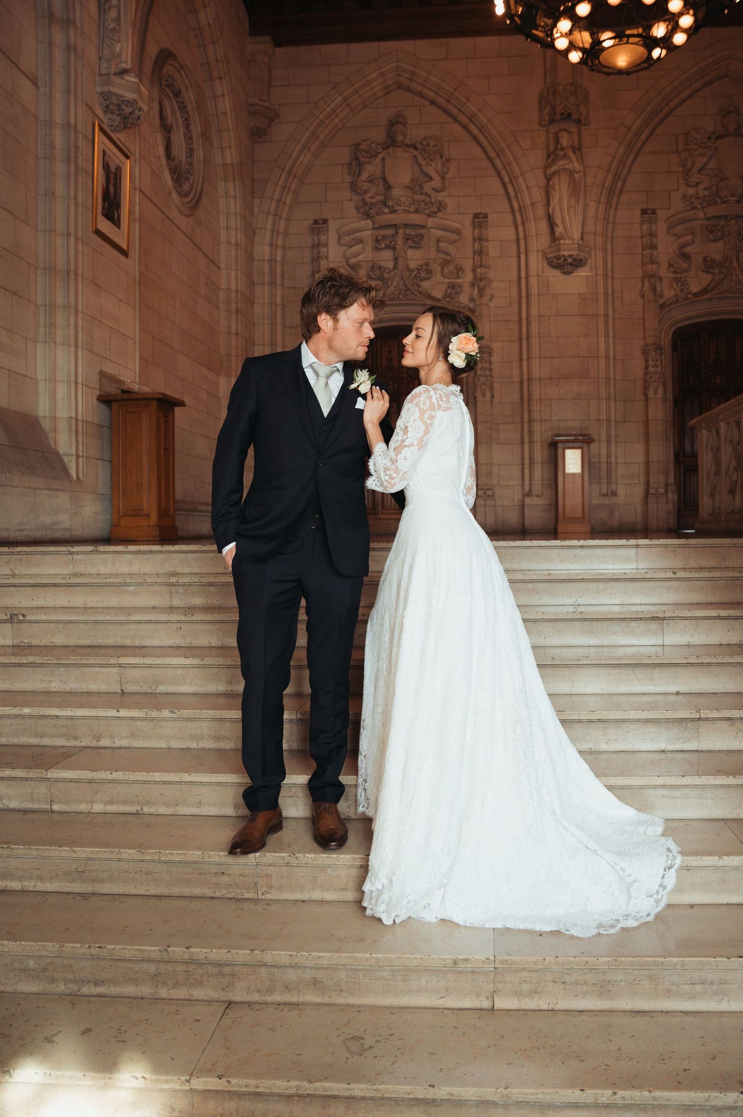 Bride and groom standing on grand stone staircase in elegant venue, sharing a romantic moment in wedding attire.