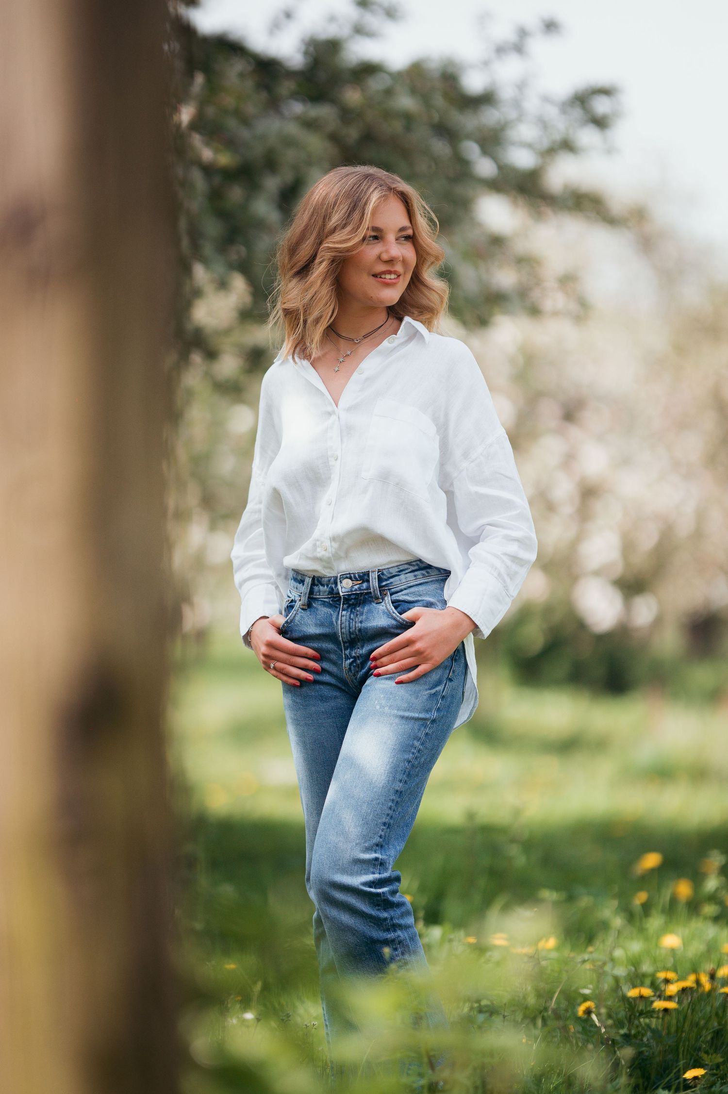 Blonde woman in white shirt and blue jeans standing outdoors among green grass and blooming trees in spring.