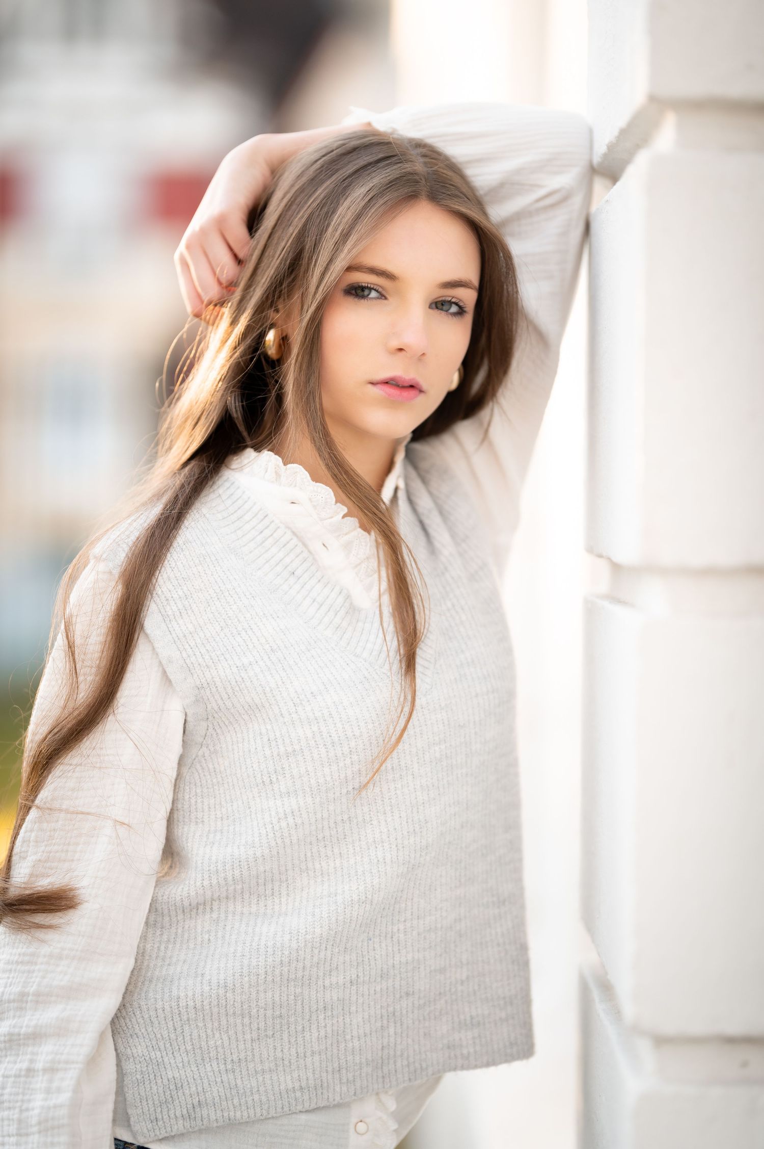 Young woman with long brown hair in a cream knit sweater posing gracefully against a white pillar outdoors.