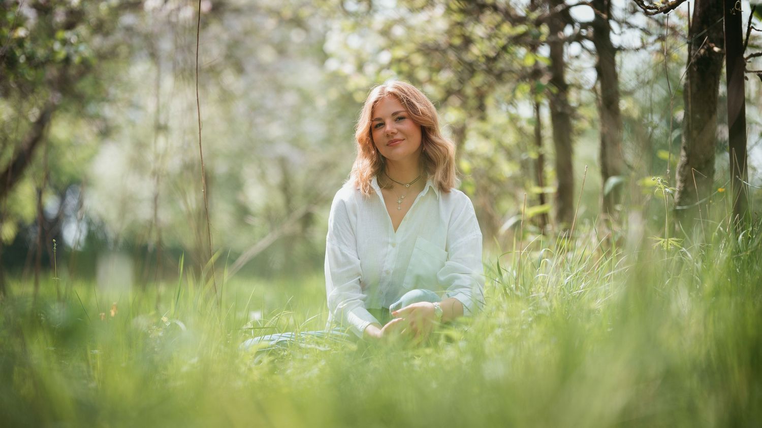 A smiling blonde woman sits peacefully in lush green grass beneath blooming spring trees in a serene outdoor setting.