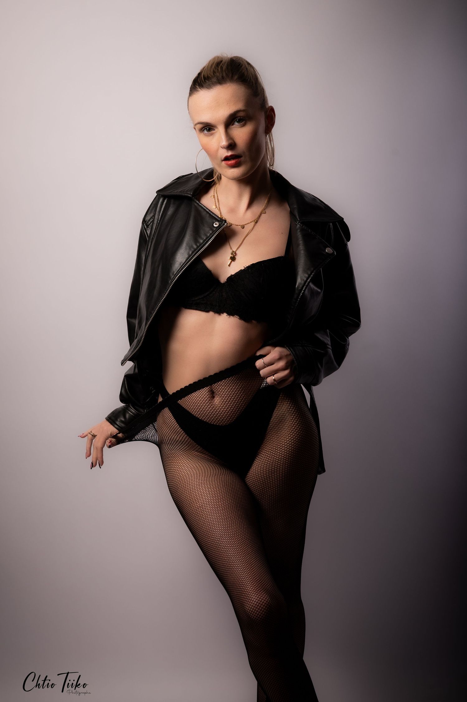 Woman posing in black lingerie, fishnet tights, and leather jacket against a neutral studio background.