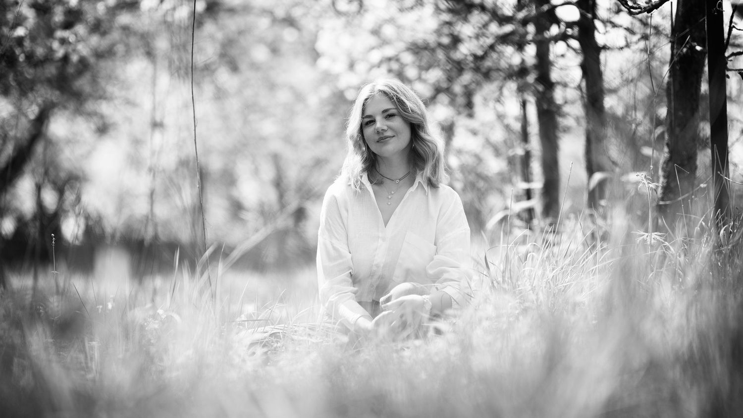 Smiling blonde woman sitting peacefully in a sunlit meadow in black and white.