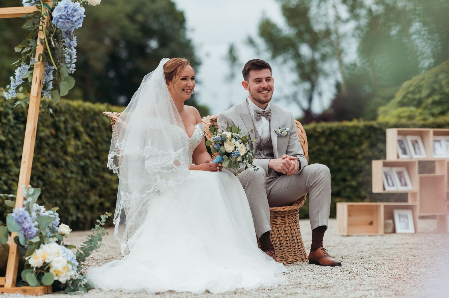 Smiling bride and groom sitting together at outdoor wedding ceremony with blue floral arch decoration.