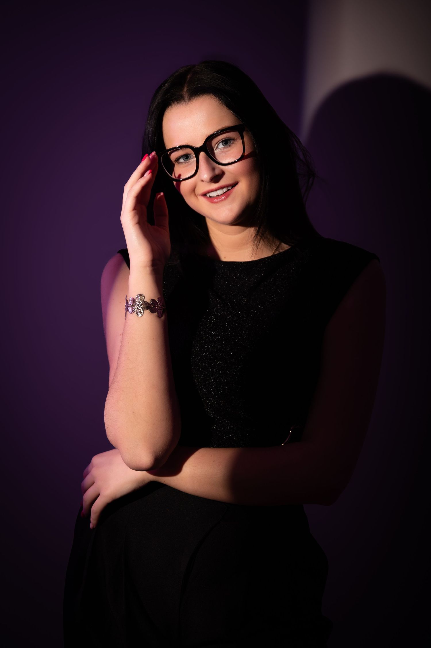A brunette woman wearing black-rimmed glasses and a black dress smiles in a purple-lit studio portrait.