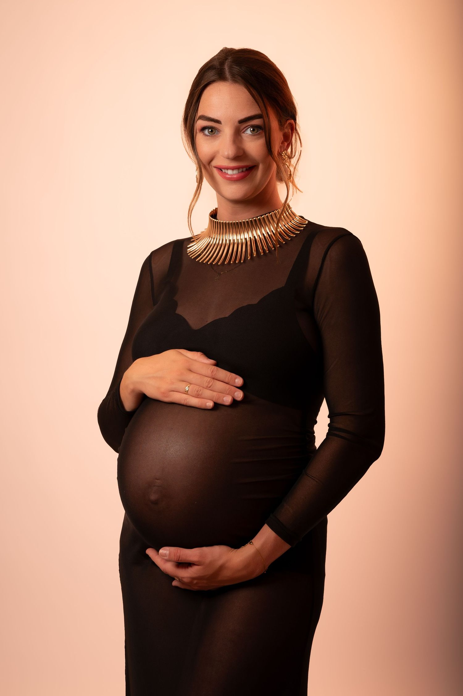 Smiling pregnant woman in black mesh dress and gold necklace cradling her baby bump against a neutral background.