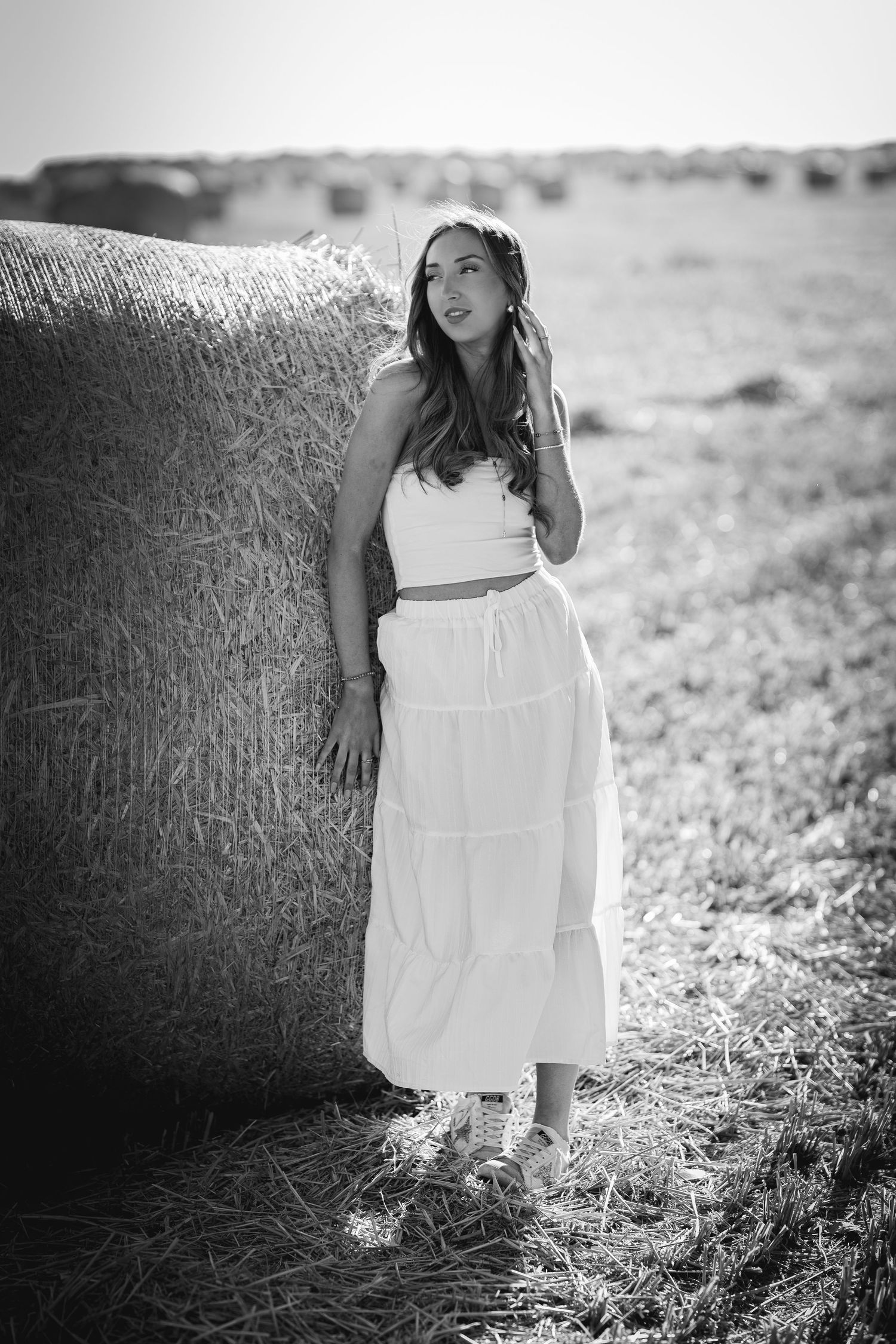 Black and white photo of a young woman in white crop top and maxi skirt leaning against a hay bale in a field.