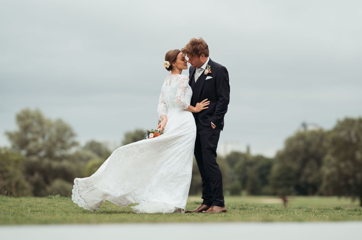 A bride and groom share a romantic embrace outdoors on their wedding day, her white gown flowing in the breeze.