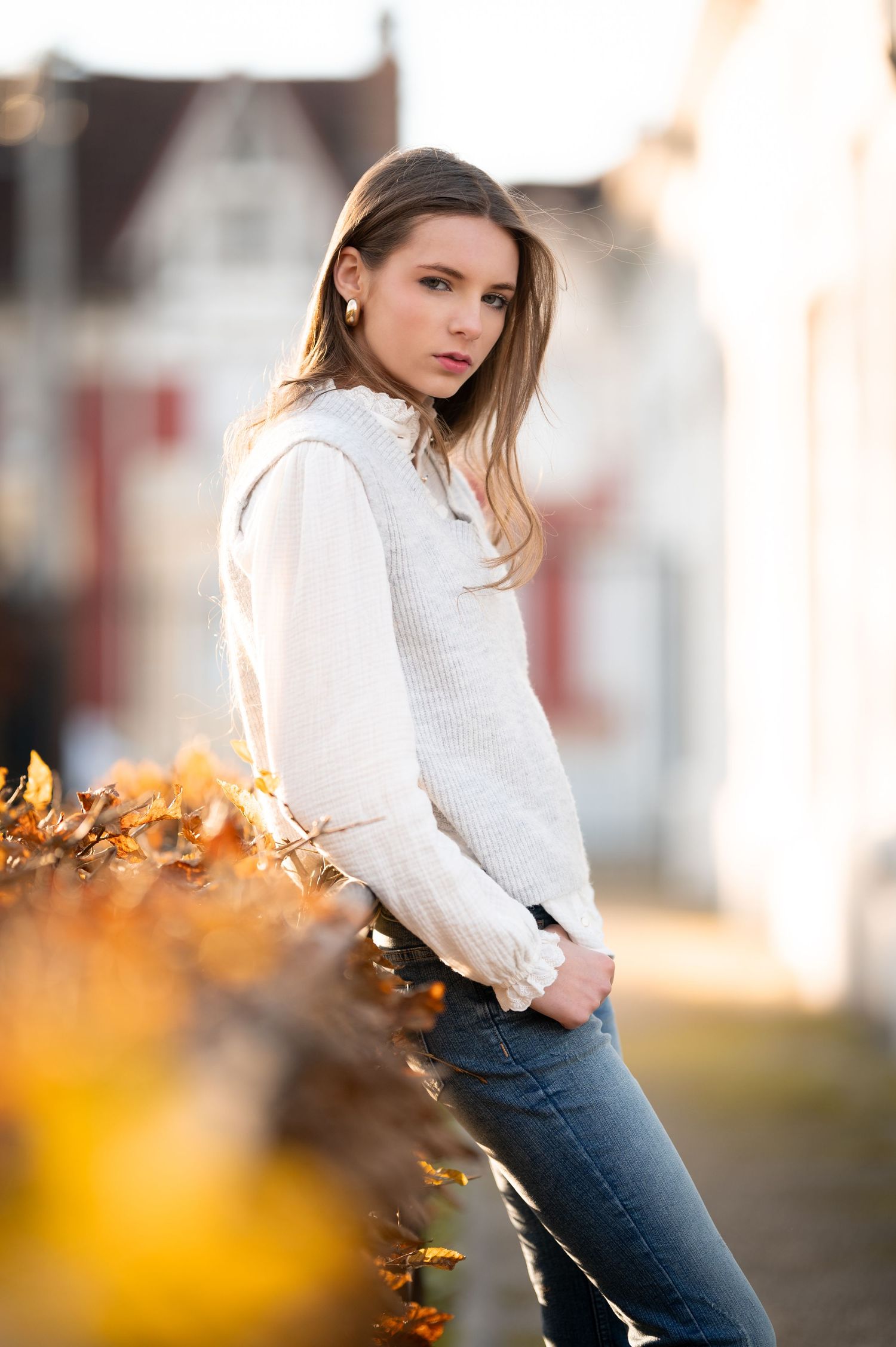 Young woman in white sweater and jeans poses outdoors among autumn leaves with blurred buildings behind her.