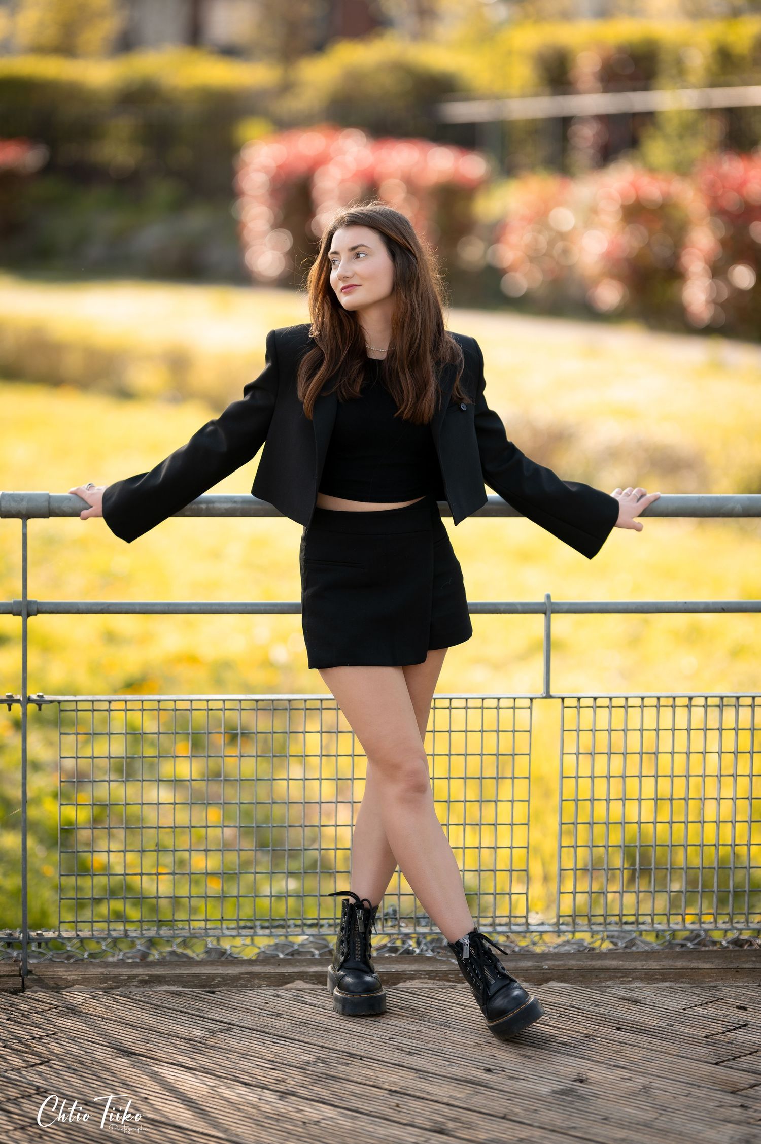 Young woman in black crop jacket, mini skirt, and combat boots posing confidently against a metal railing outdoors.