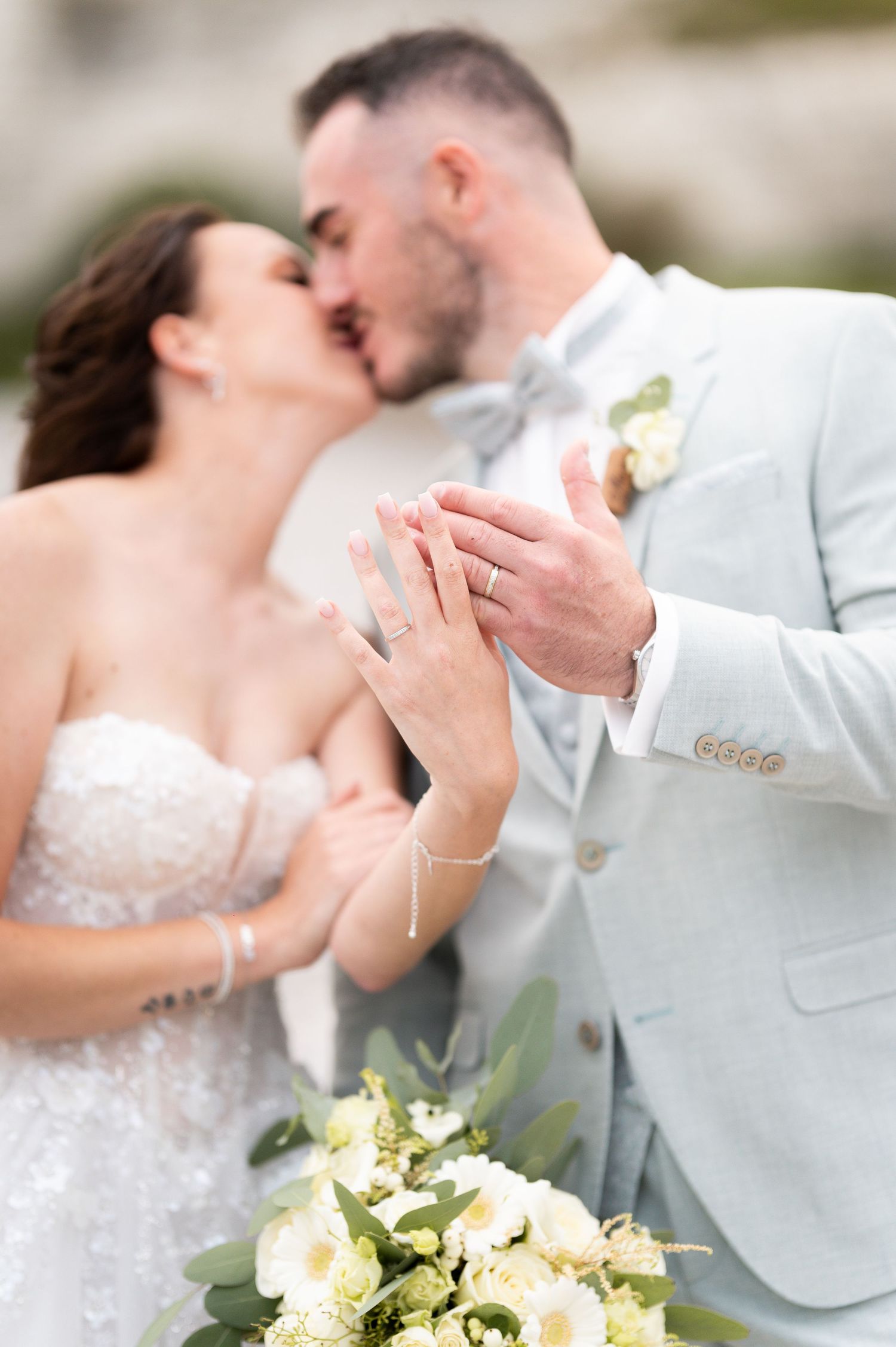 Bride and groom kissing while displaying wedding rings, with white floral bouquet in foreground.