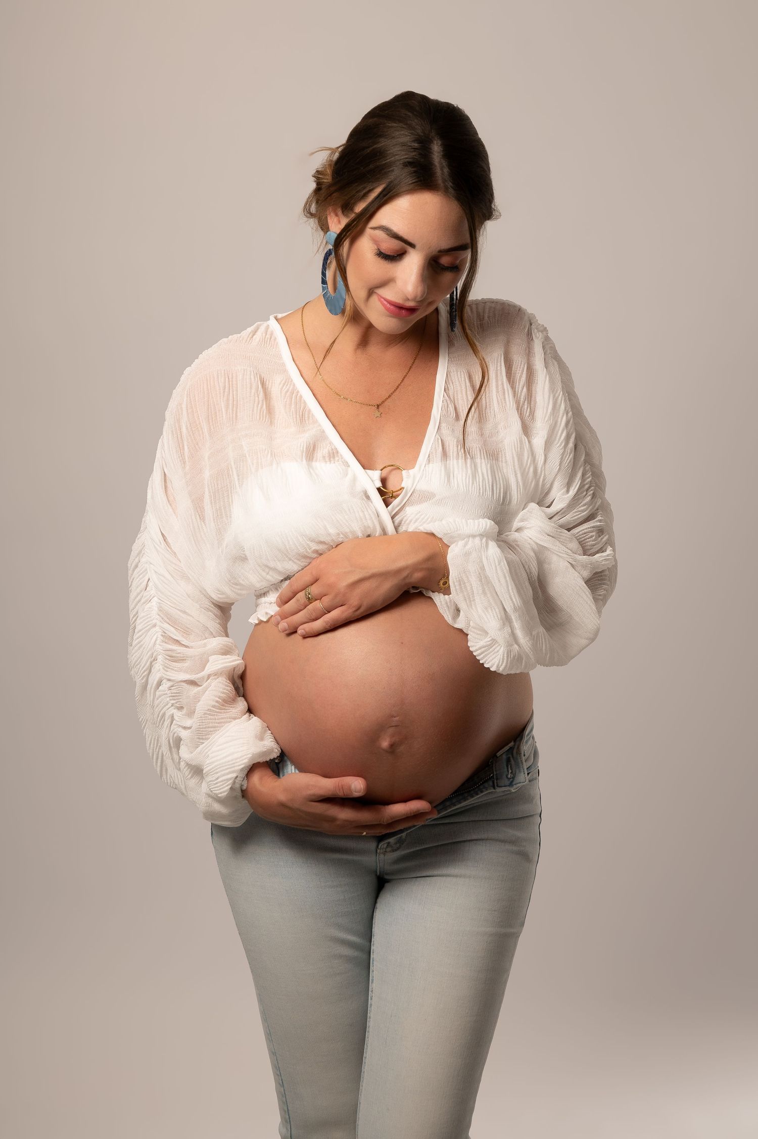 Pregnant woman in white blouse and jeans cradling her baby bump, smiling down, wearing blue earrings and a necklace.