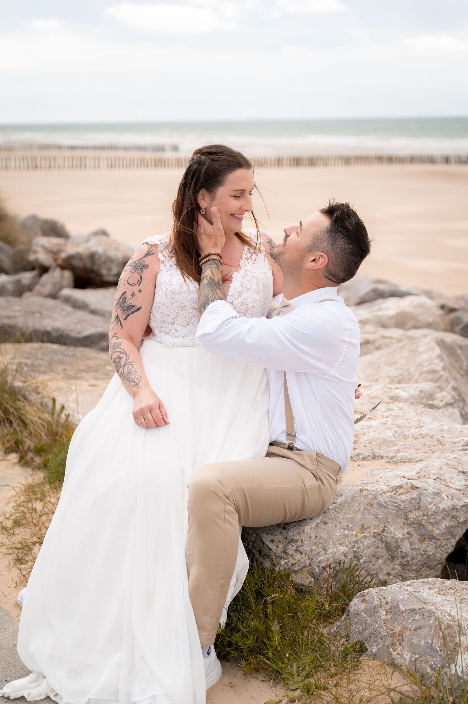 Groom gazes lovingly at bride seated on rocks with ocean beach backdrop.