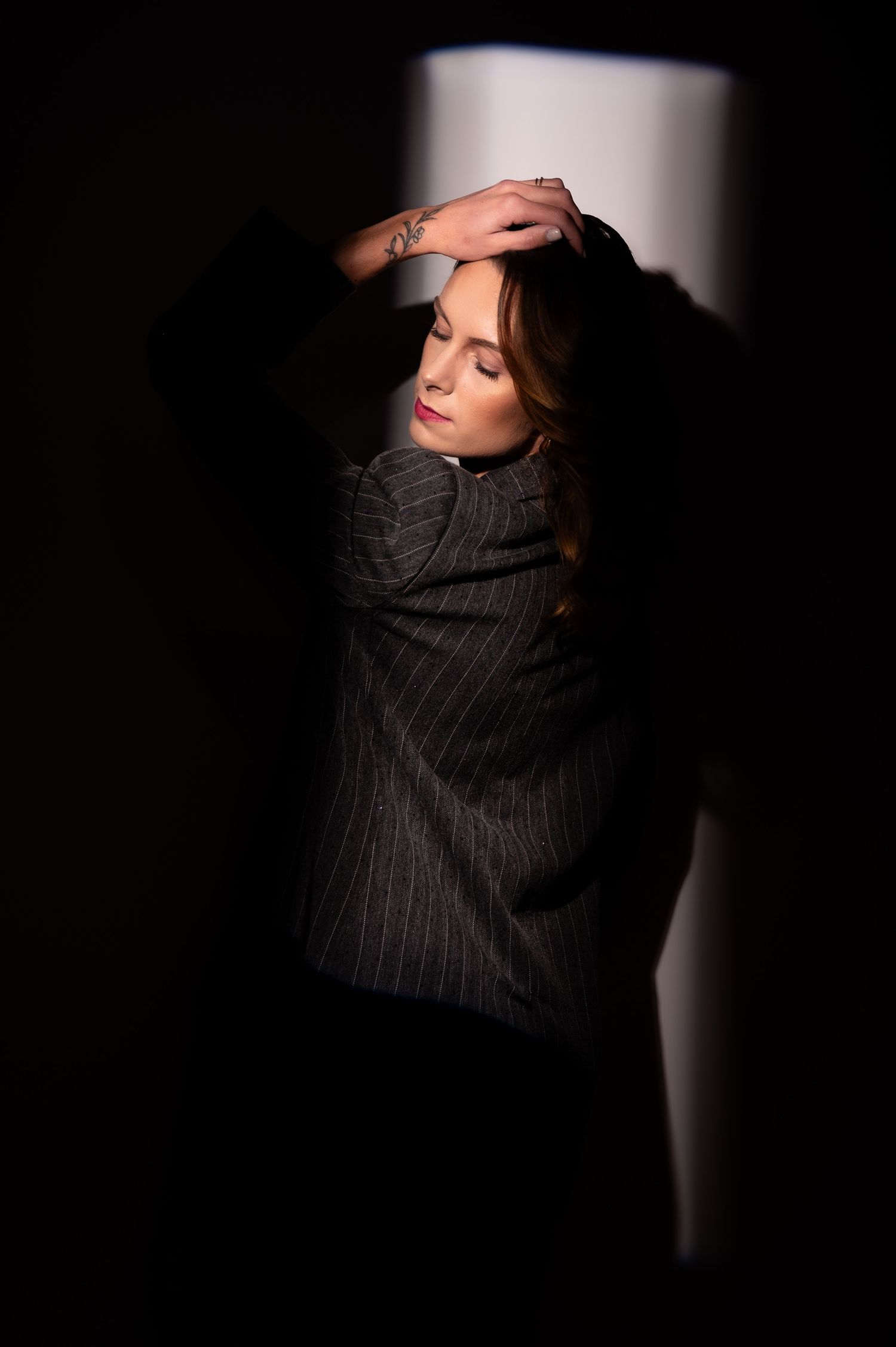 A woman in a striped blazer poses dramatically against a wall in striking light and shadow.