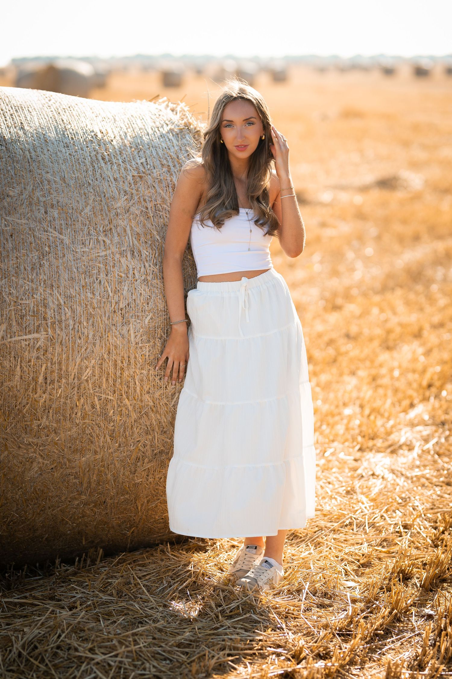 Woman in white crop top and maxi skirt posing beside a hay bale in a golden sunlit field.