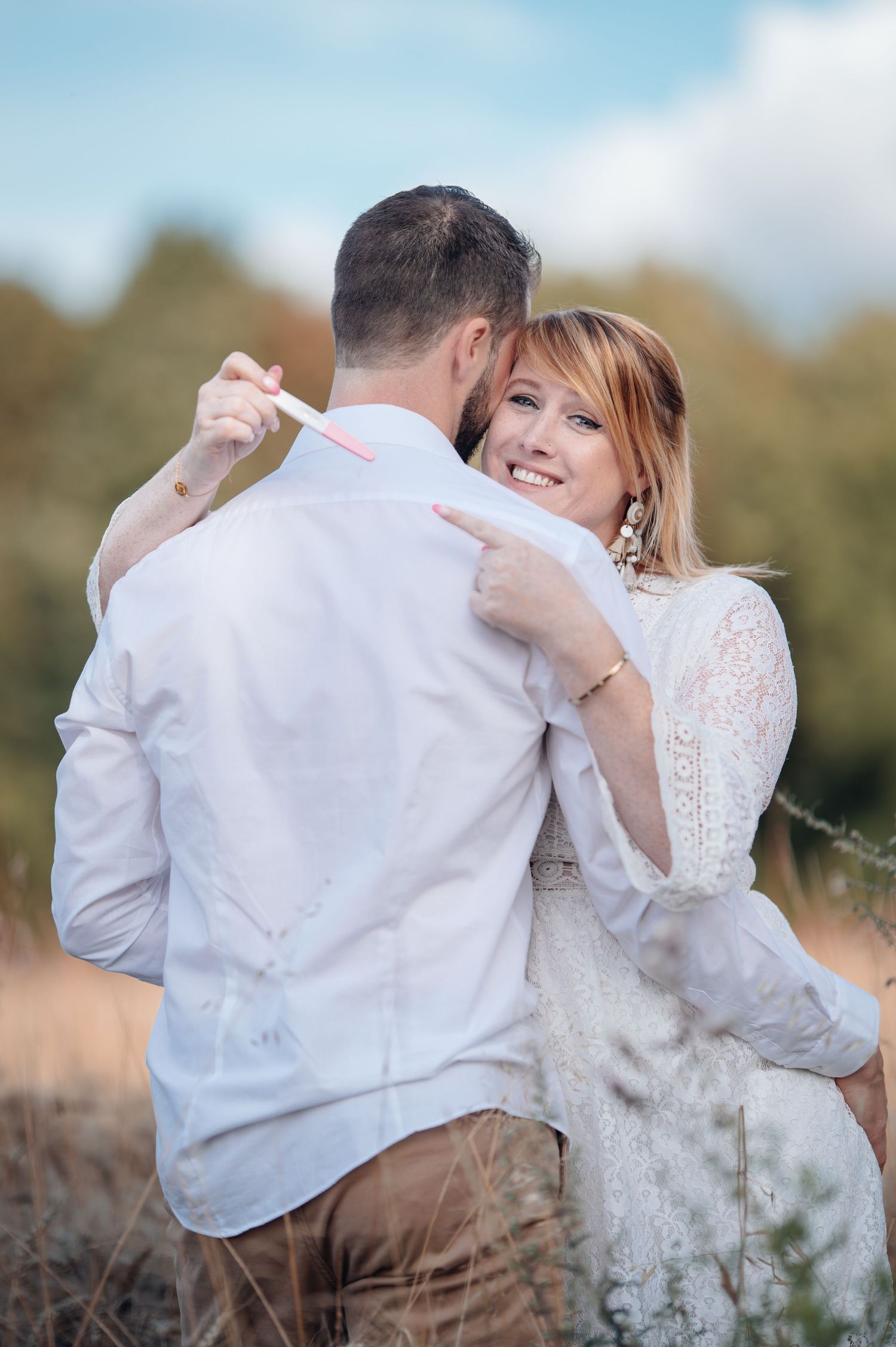 A smiling woman in a white lace dress embraces a man in a white shirt outdoors in a field.