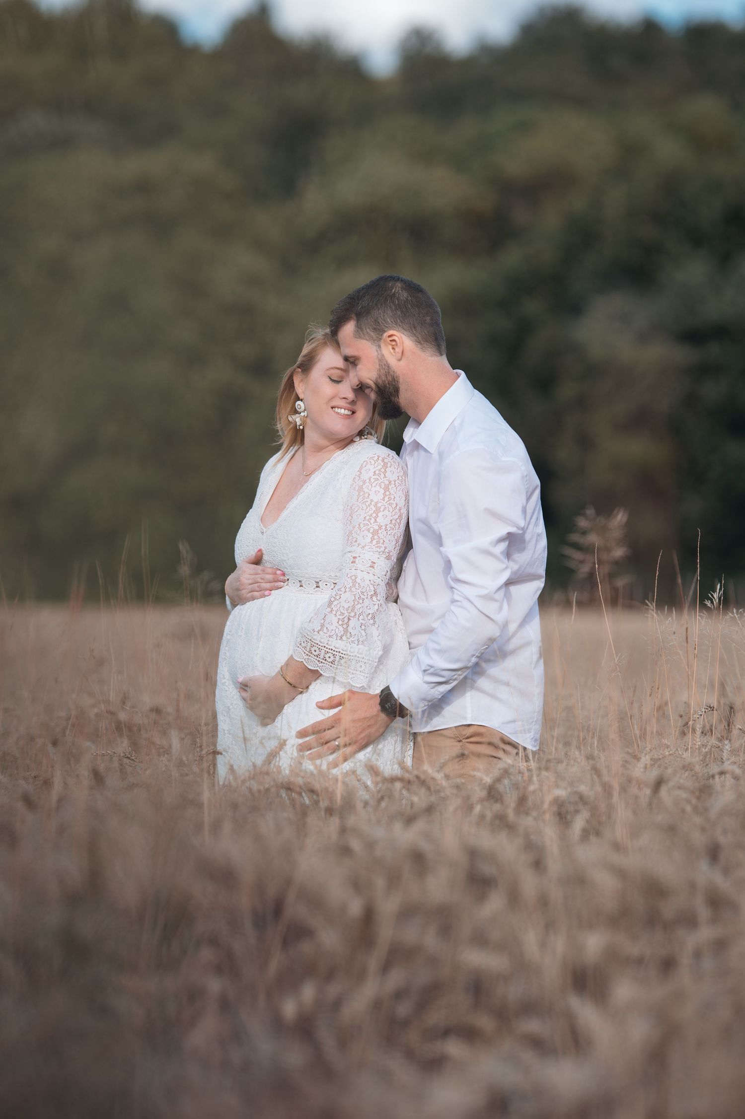 Pregnant couple embracing in golden field, woman in white lace dress, man in white shirt, with trees in background.