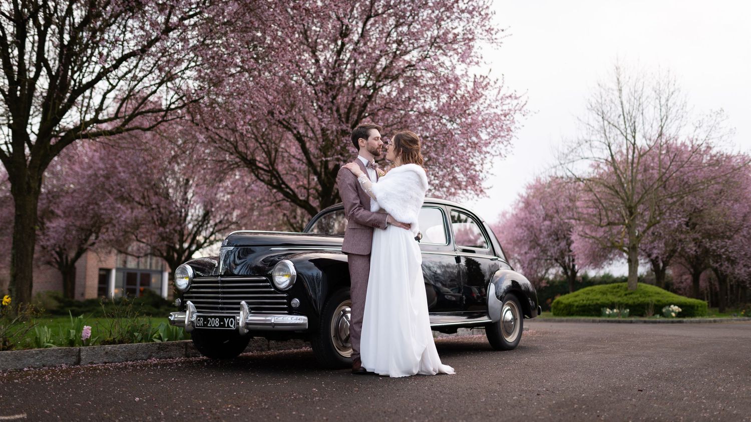 Bride and groom embracing beside a classic black vintage car under blooming pink cherry blossom trees.