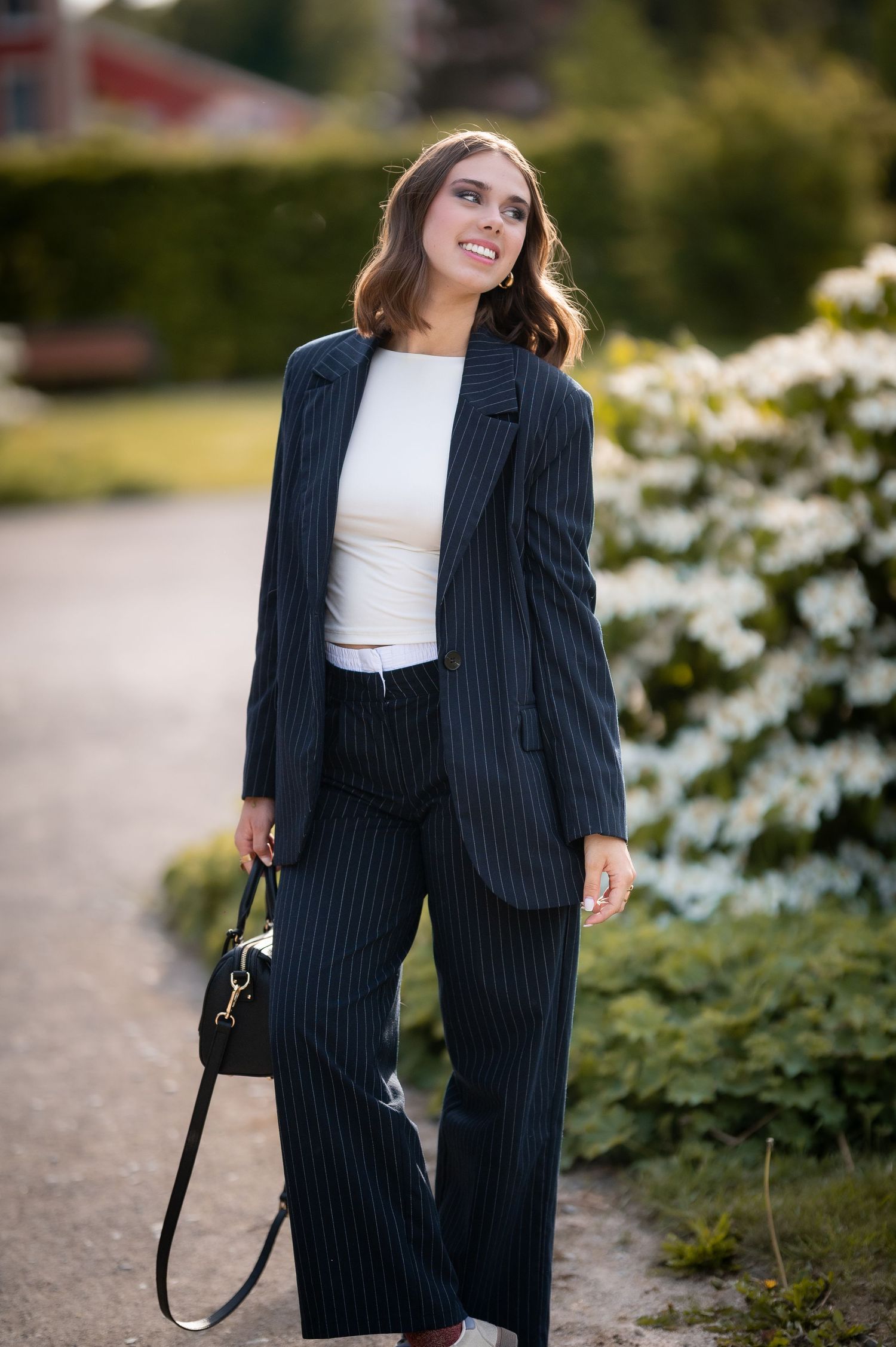 Woman in navy pinstripe suit with white top and black handbag smiling outdoors near blooming shrubs.