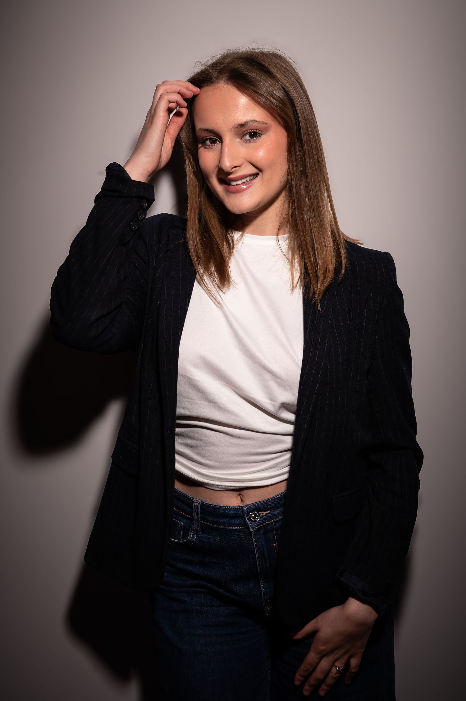 Smiling young woman in black blazer and white crop top with jeans posing casually in a studio portrait setting.