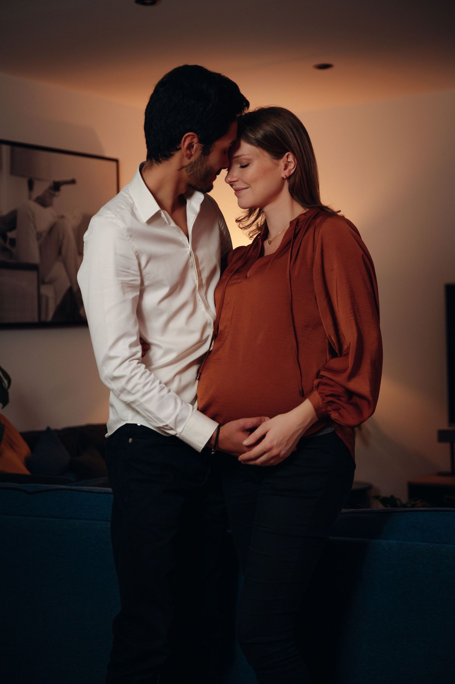 A couple shares a tender moment, the woman visibly pregnant, embracing in warm ambient home lighting.