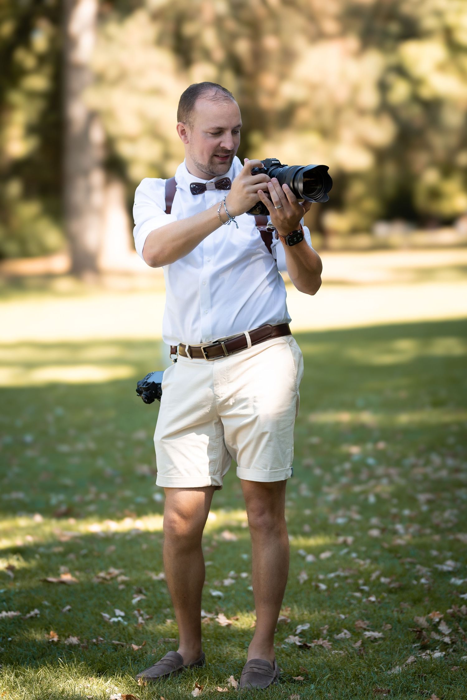 Man in white shirt and shorts holding a camera in a sunny park, reviewing photos outdoors.