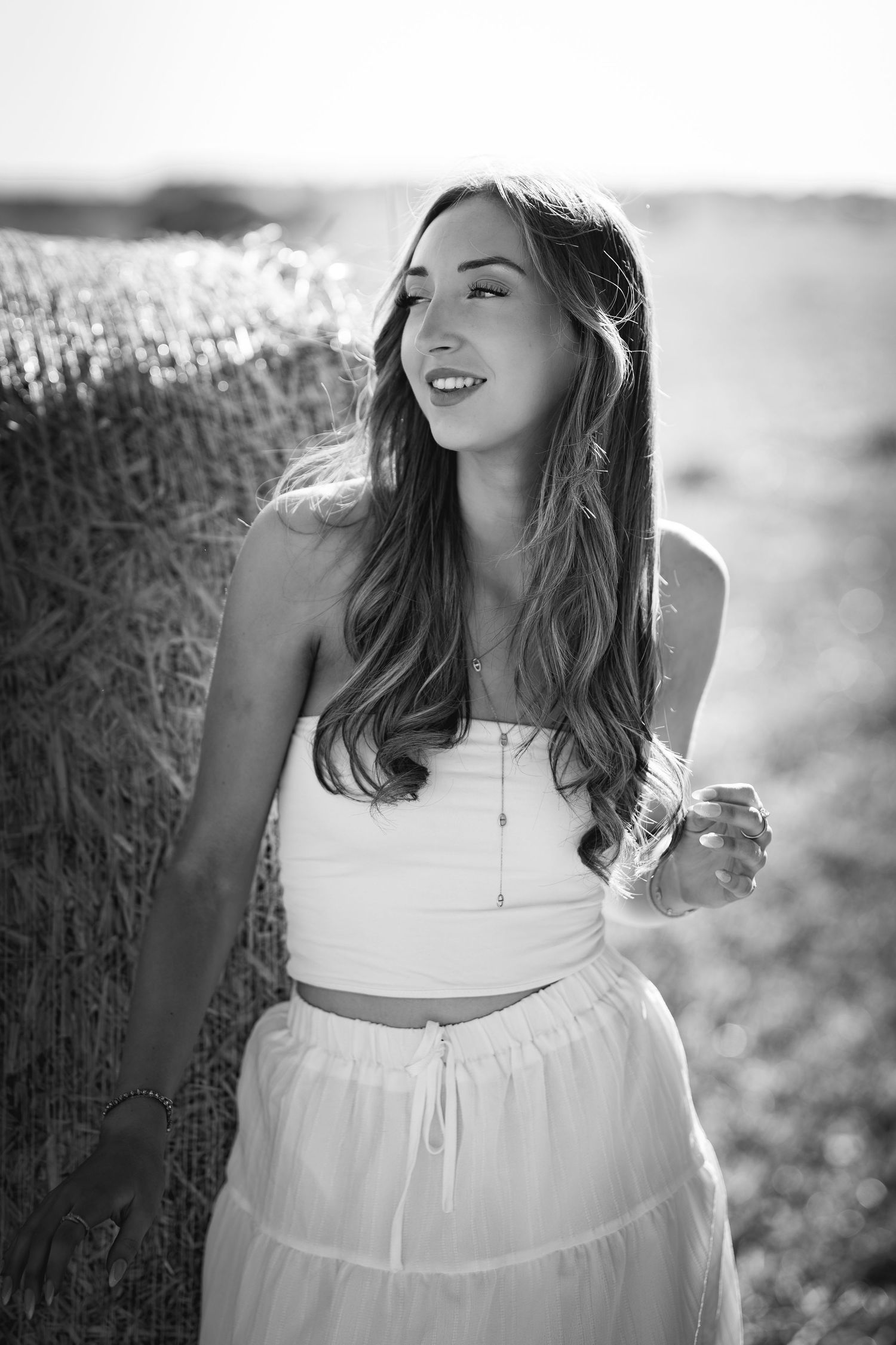 Black and white photo of a young woman in a white crop top and skirt leaning against a hay bale in a field.