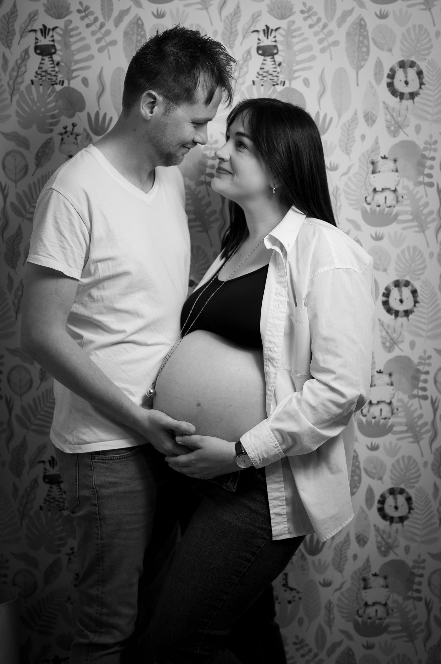Black and white photo of a couple tenderly holding a pregnant belly, standing against nursery wallpaper.