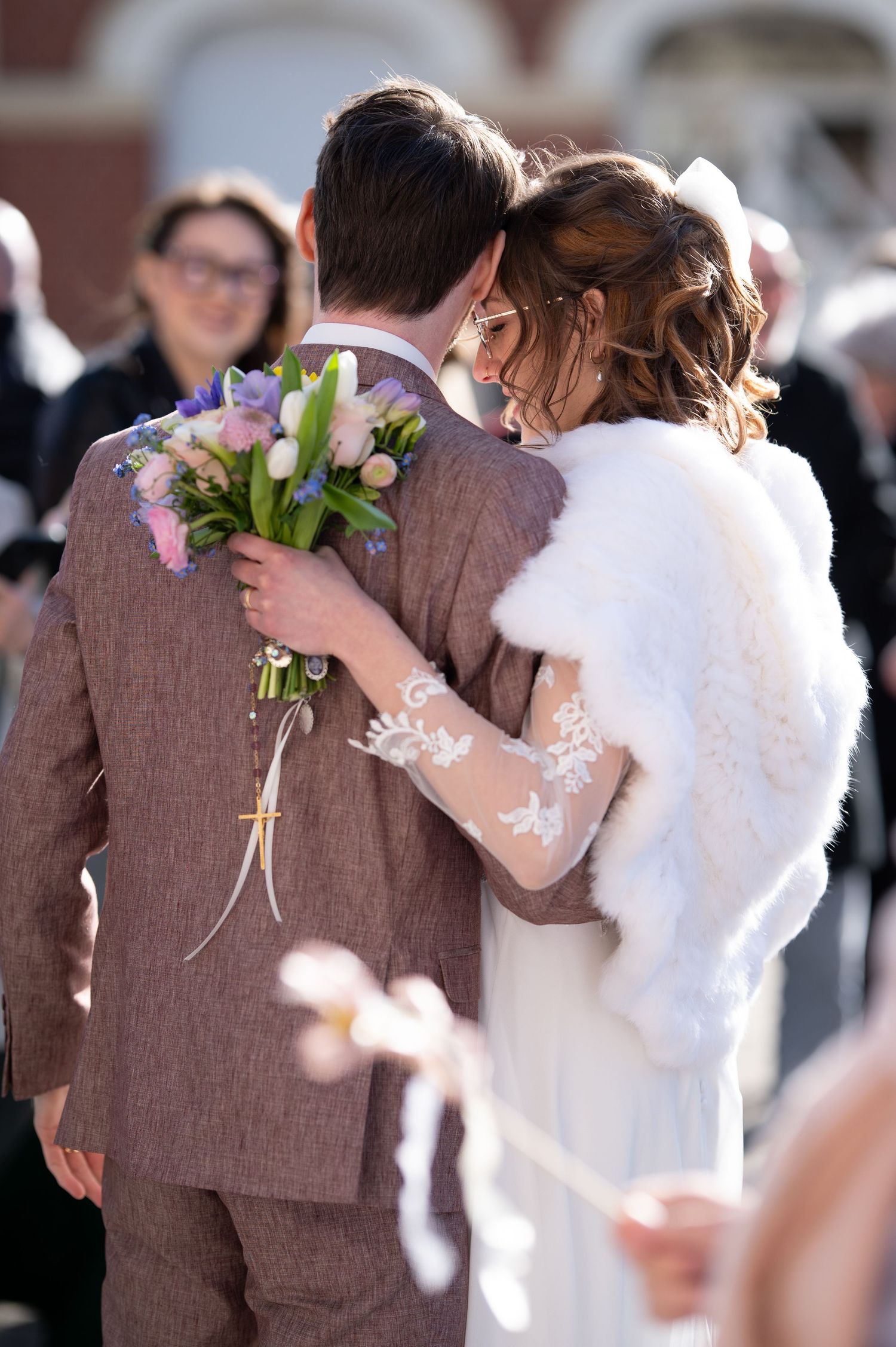 Bride in lace-sleeved gown with white fur stole embraces groom in brown suit holding pastel spring wedding bouquet.