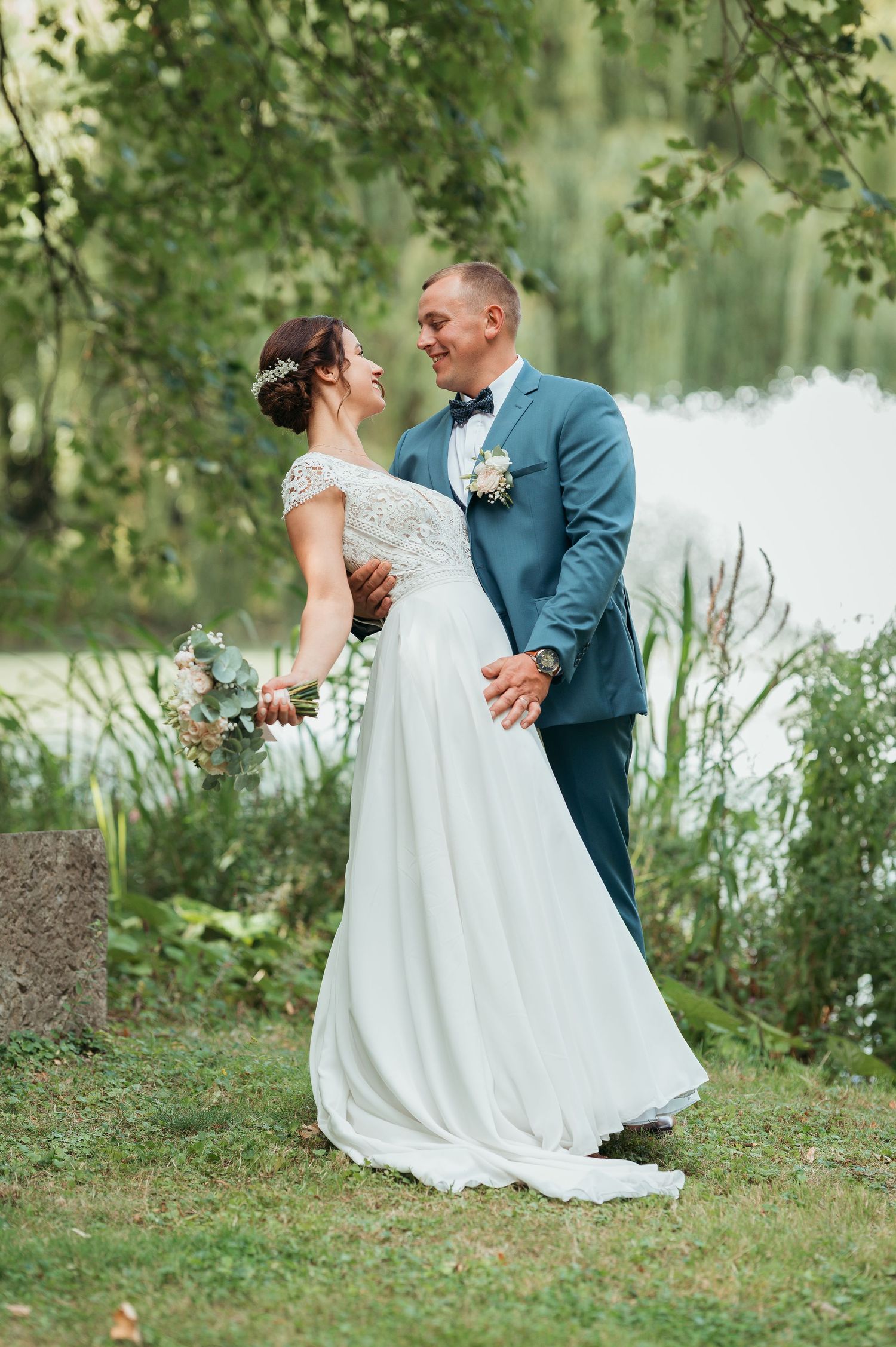A bride and groom share a romantic moment by a lakeside, she in a white gown, he in a blue suit with bow tie.