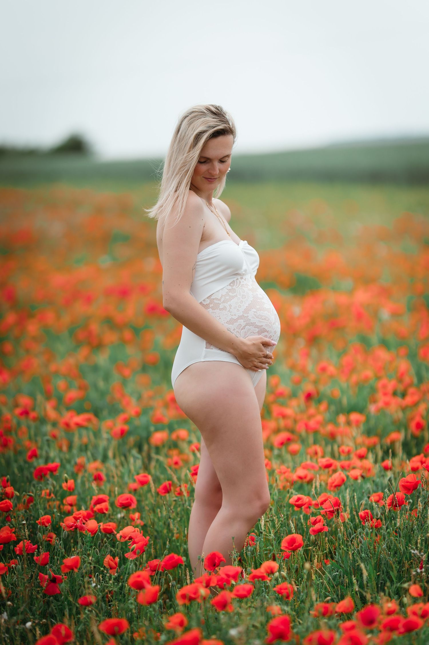 A pregnant blonde woman in a white bodysuit cradles her belly in a vibrant red poppy field.