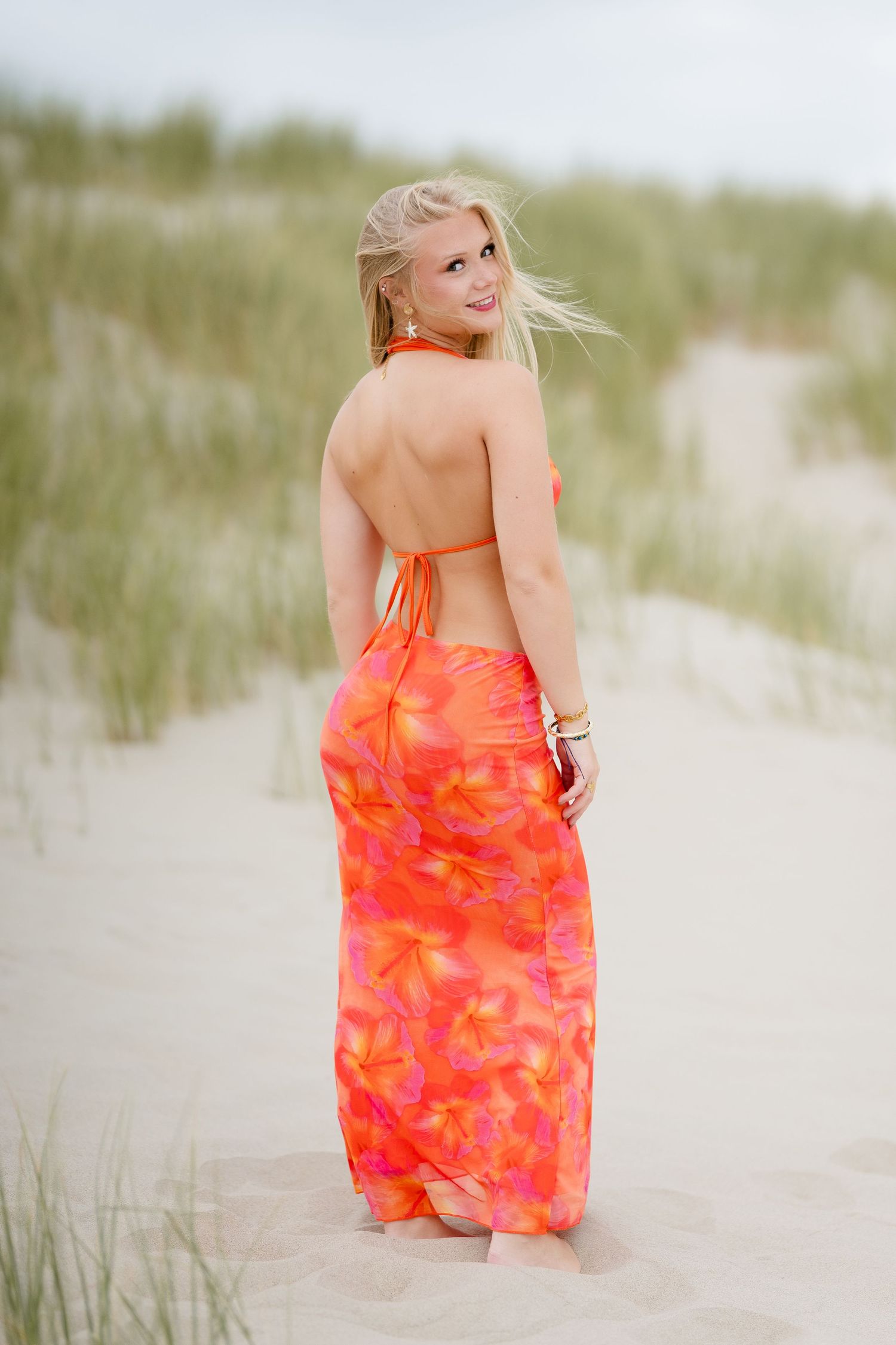 Blonde woman in orange floral halter maxi dress standing on sandy beach dunes with grass in background.