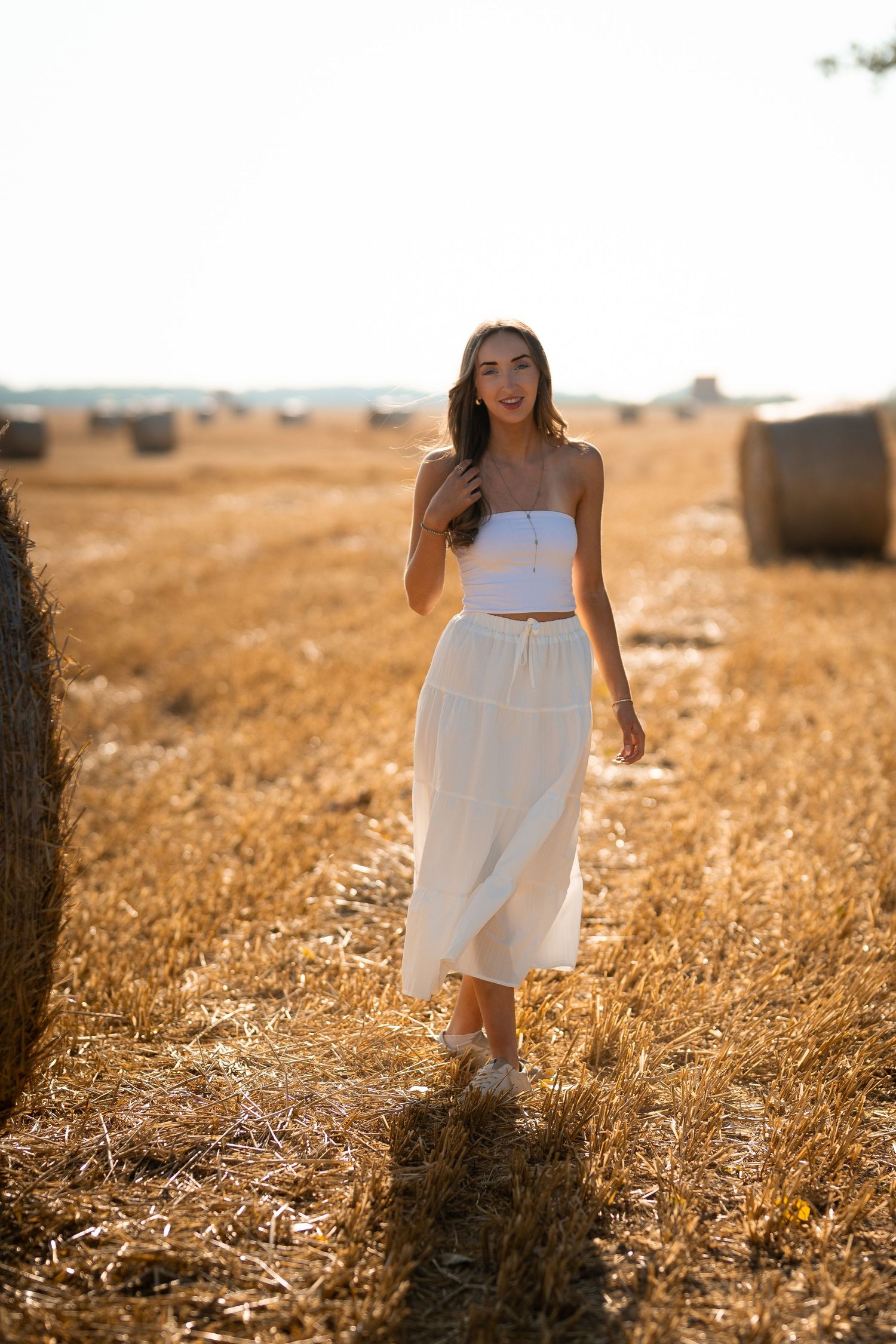 Young woman in white crop top and midi skirt posing in golden hay field with round bales at sunset.