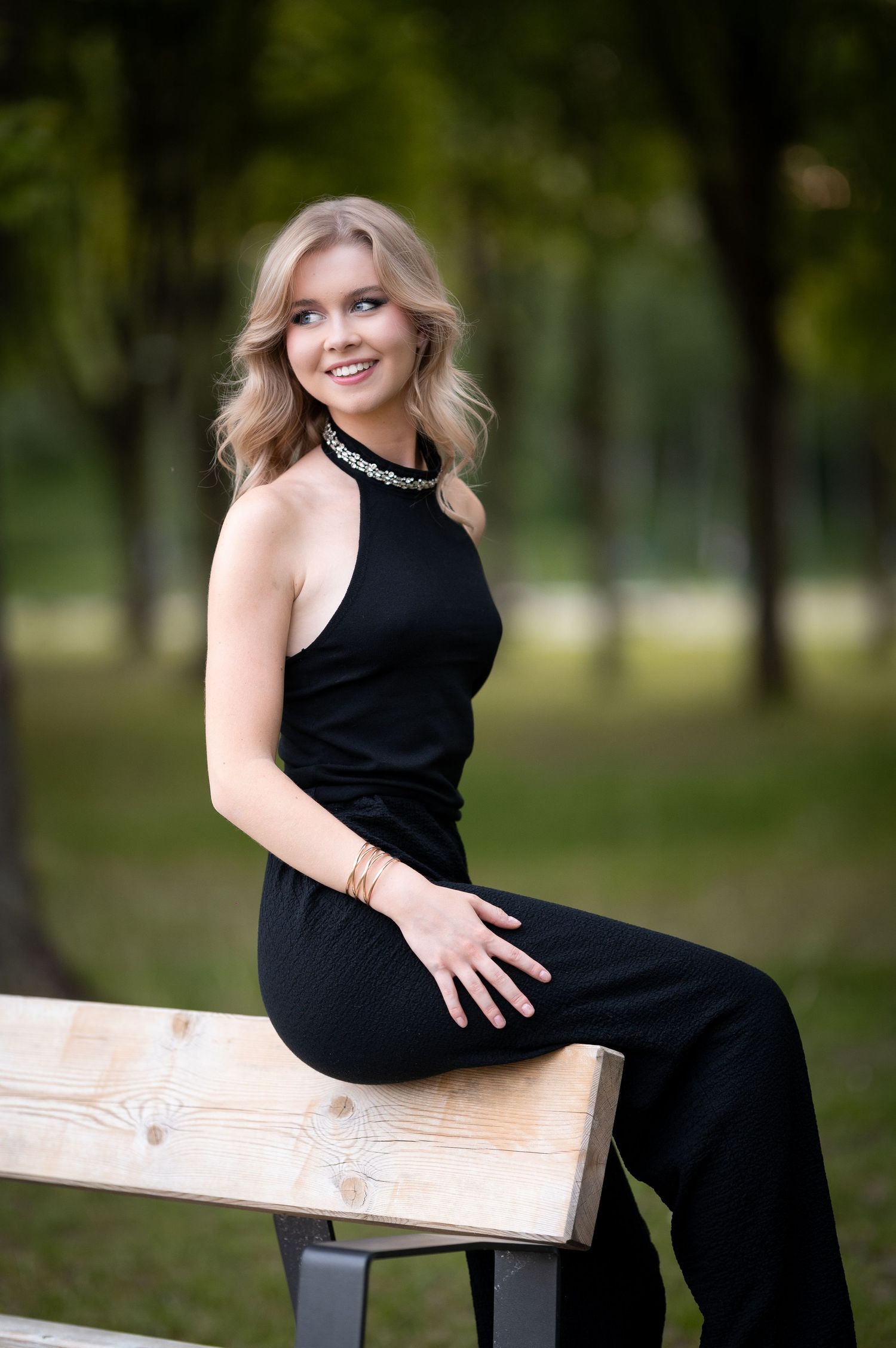 Smiling blonde woman in black halter jumpsuit sitting on a wooden bench in a lush green park.