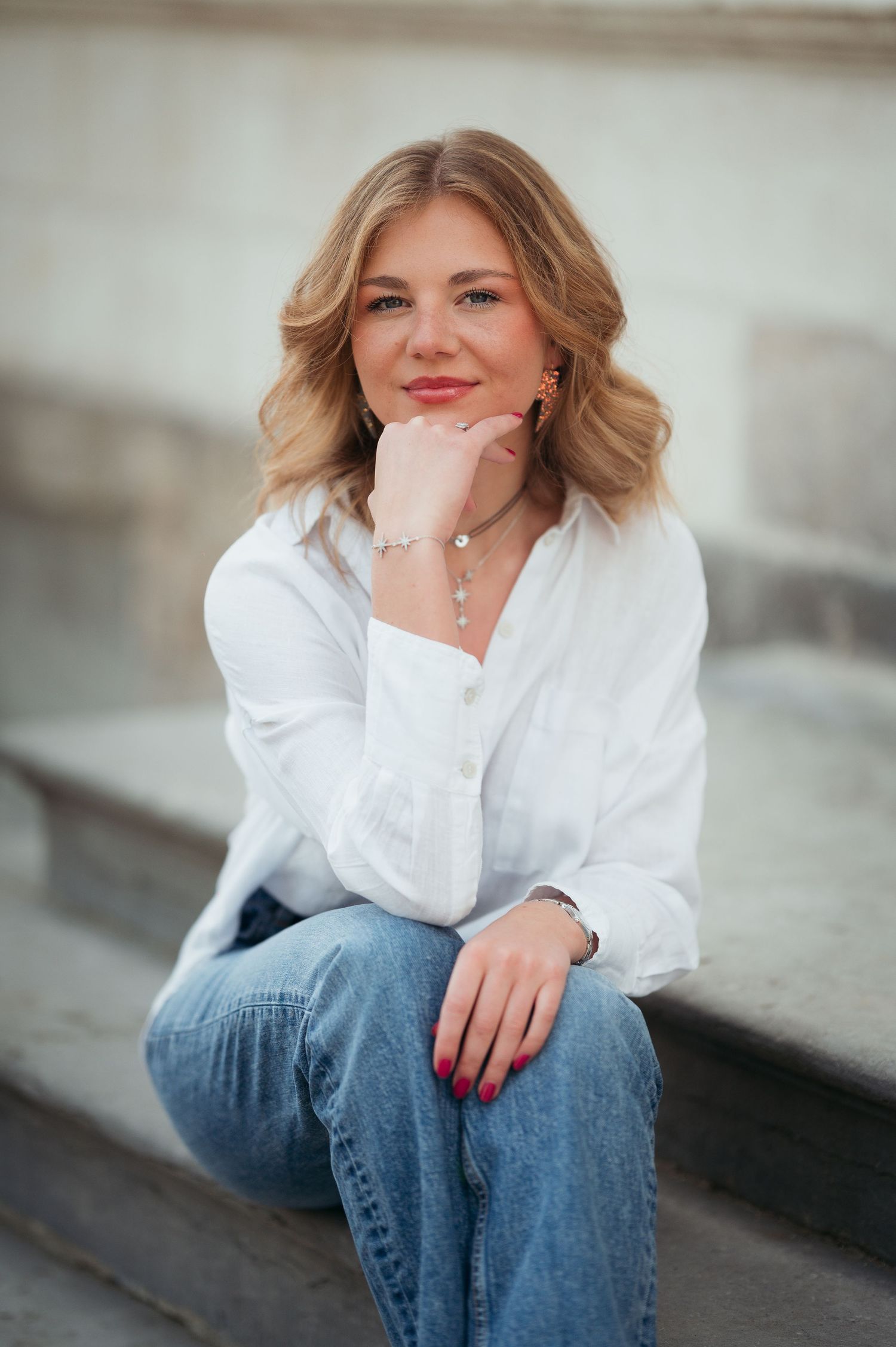 Young blonde woman in white shirt and jeans sitting on steps, smiling with hand on chin, wearing bracelet.