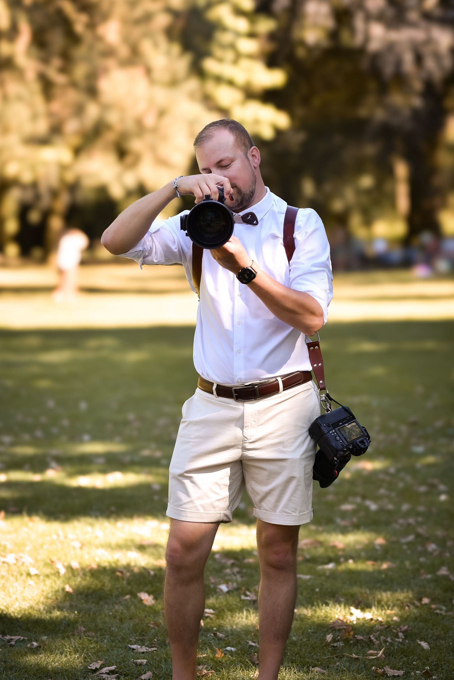 A male photographer in white shirt and khaki shorts takes photos with a DSLR camera in a sunny park.
