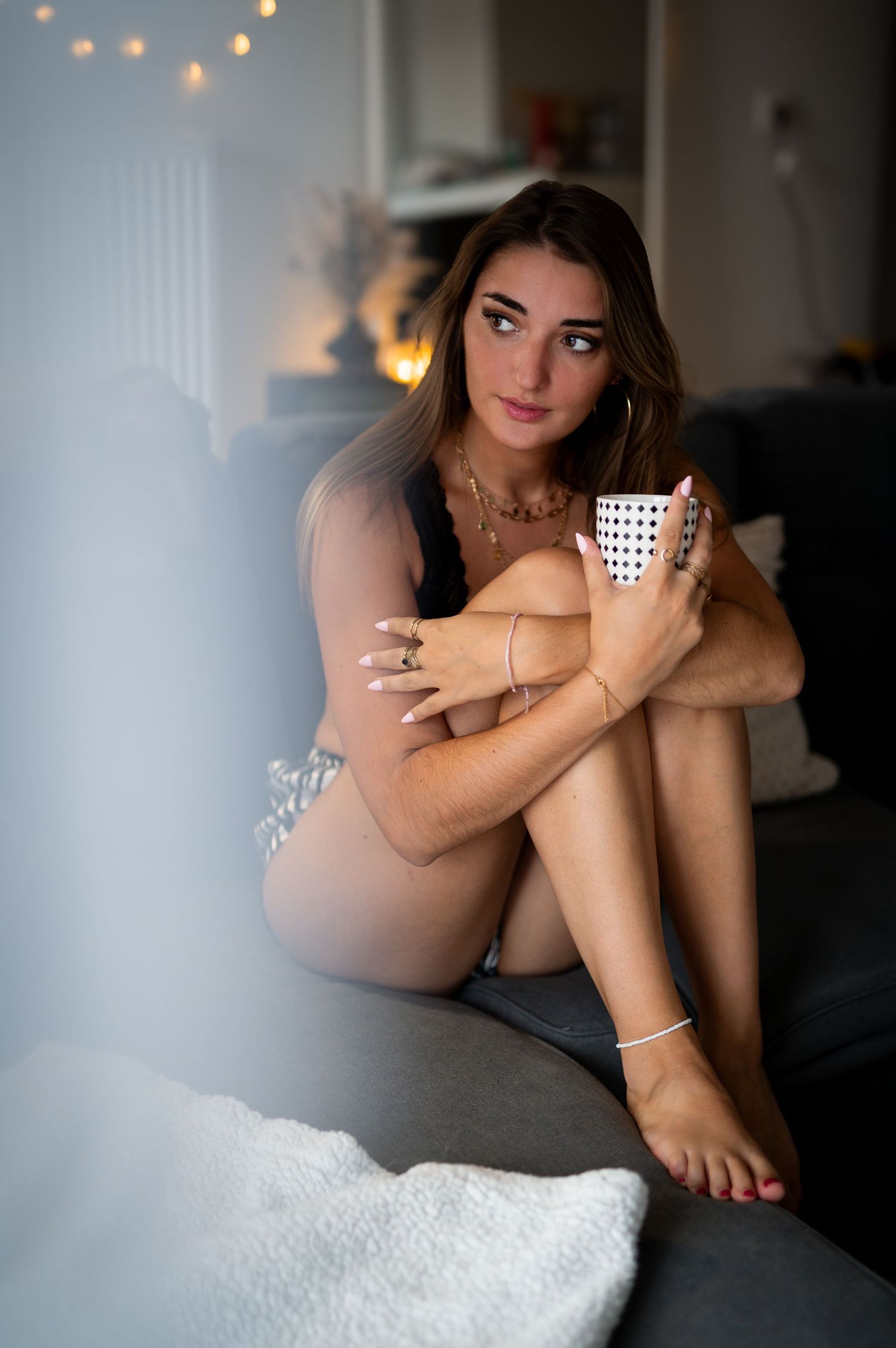 Young woman in lingerie sitting on couch hugging knees, holding a mug, with warm bokeh lights in background.