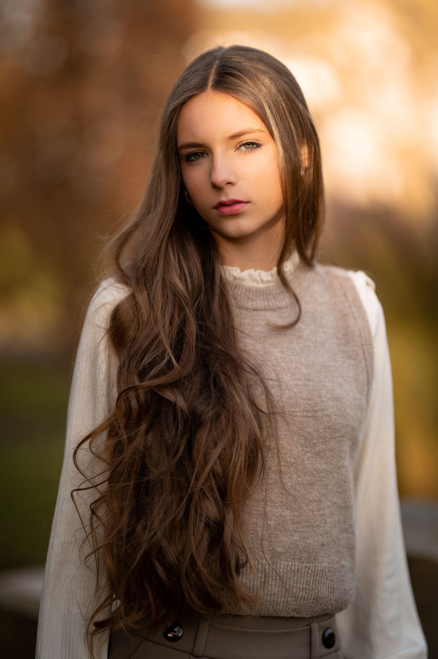 Young woman with long wavy brown hair wearing a beige sweater, posing outdoors in warm autumn light.