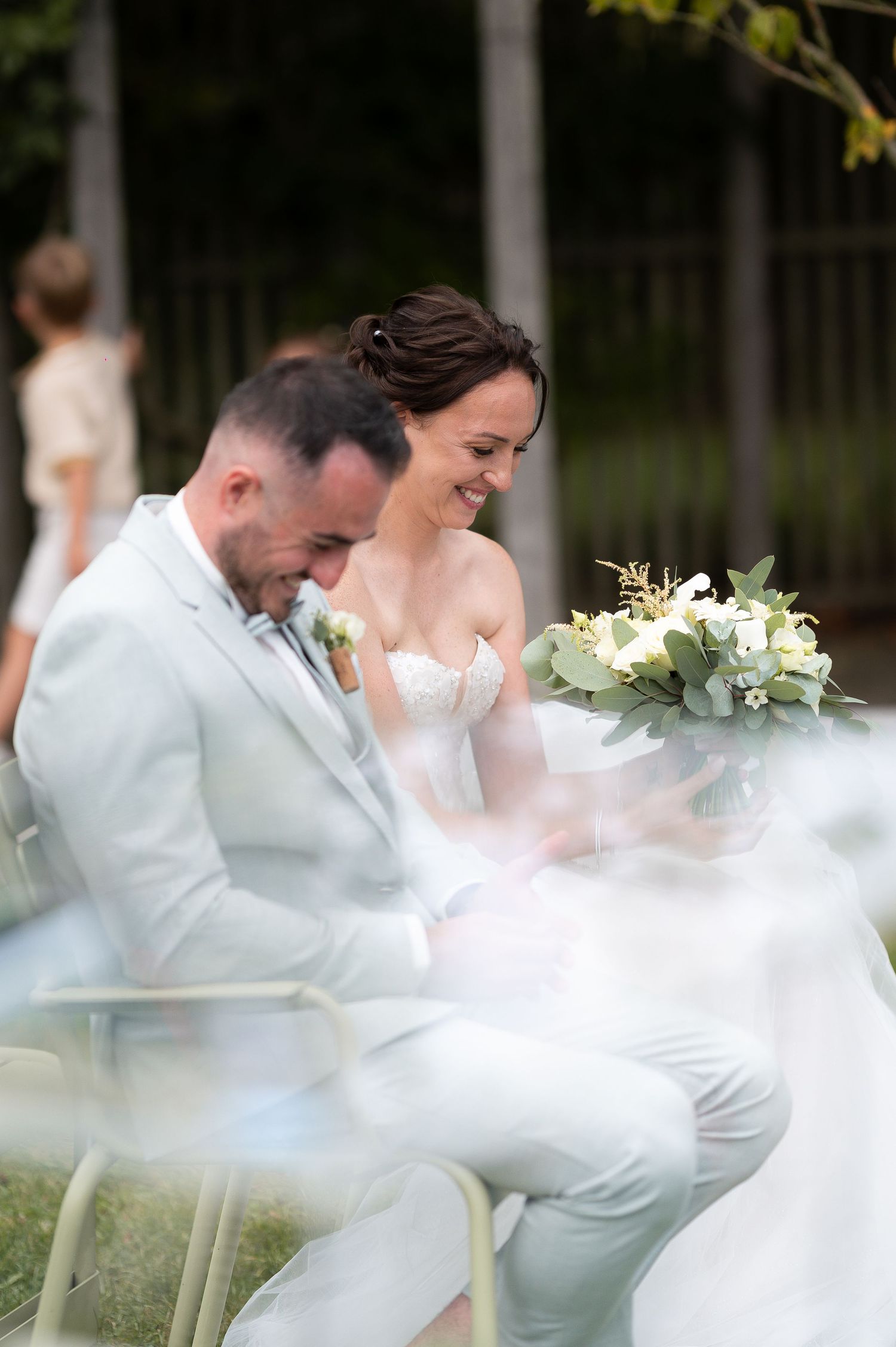 Bride and groom laughing together during outdoor wedding ceremony, bride holding white and green bouquet, groom in light gray suit.