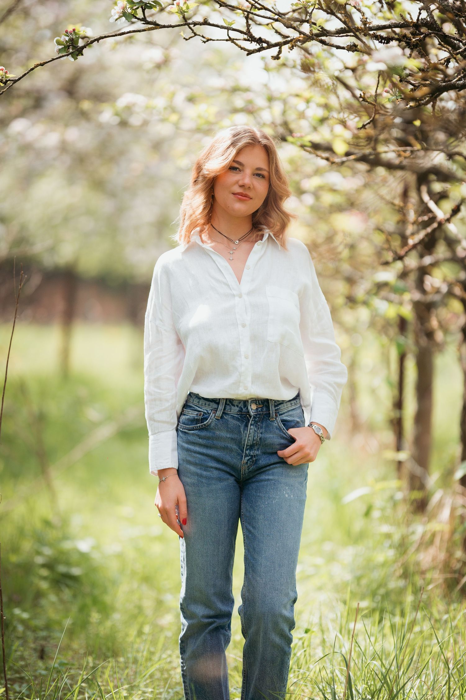 Young woman in white shirt and jeans standing in blooming spring orchard with flowering trees.