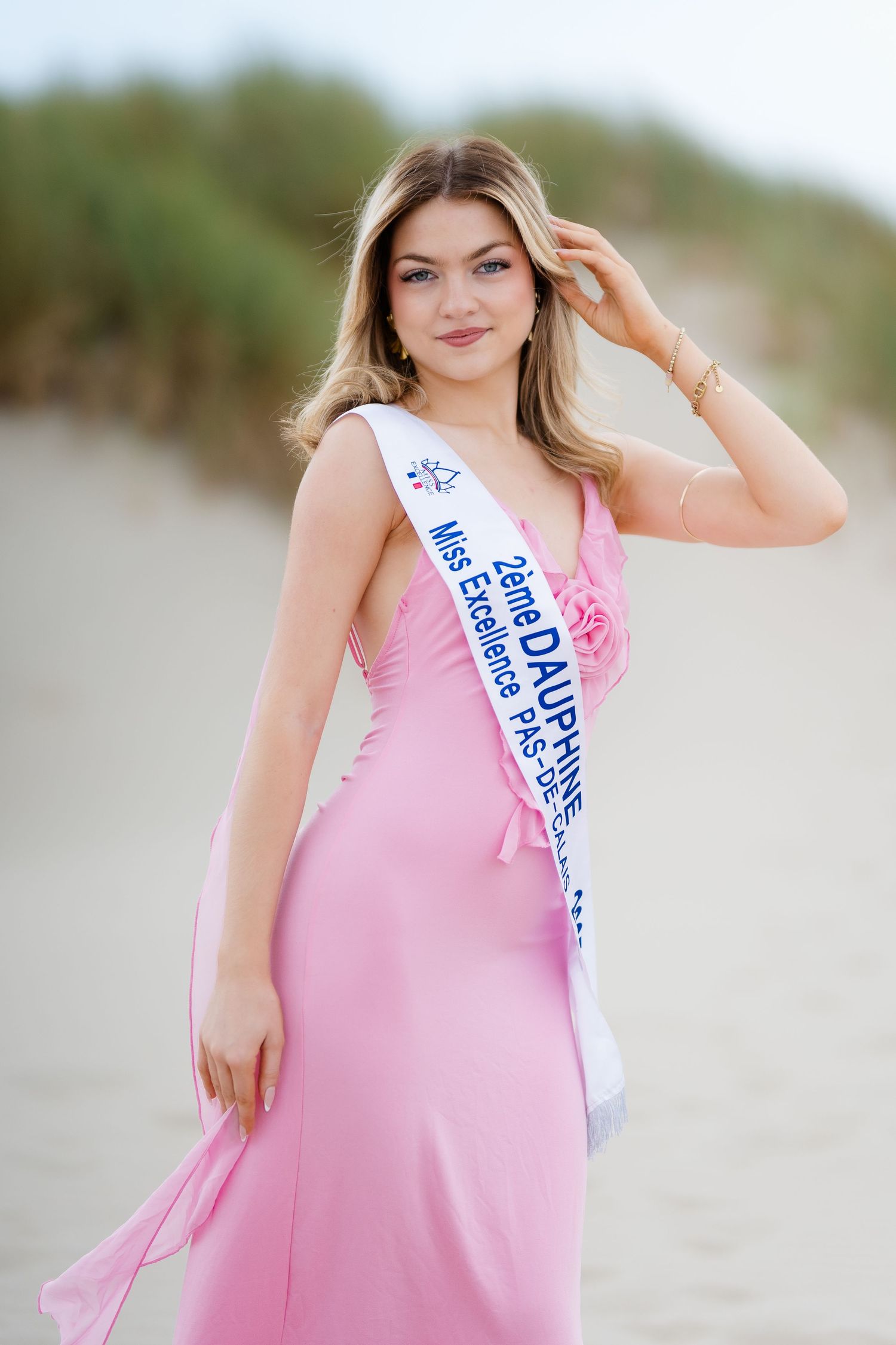 Young woman in pink gown wearing a beauty pageant sash on a sandy beach, posing gracefully outdoors.