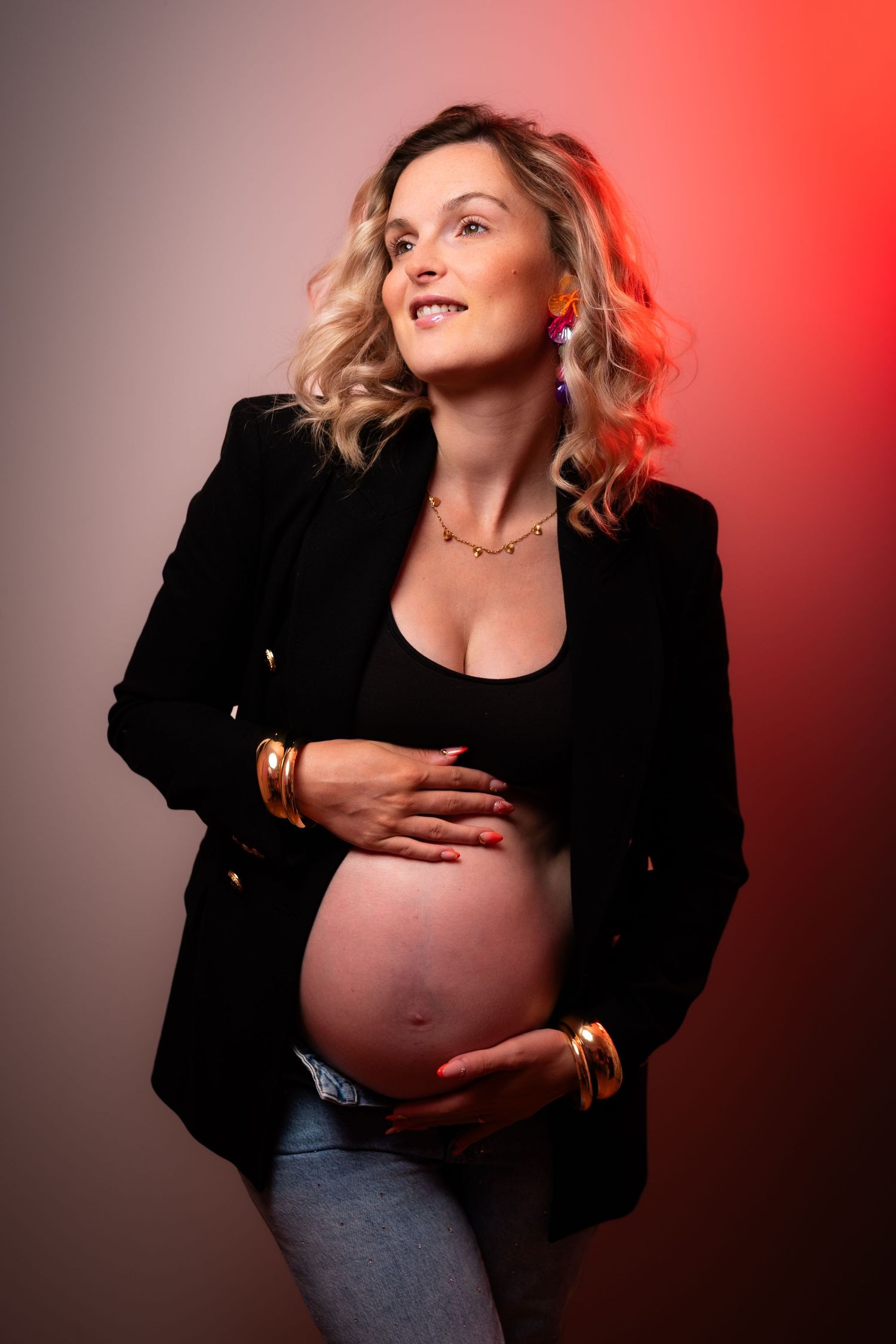 Pregnant woman in black blazer and jeans cradling baby bump, wearing gold jewelry and colorful earrings, studio portrait.