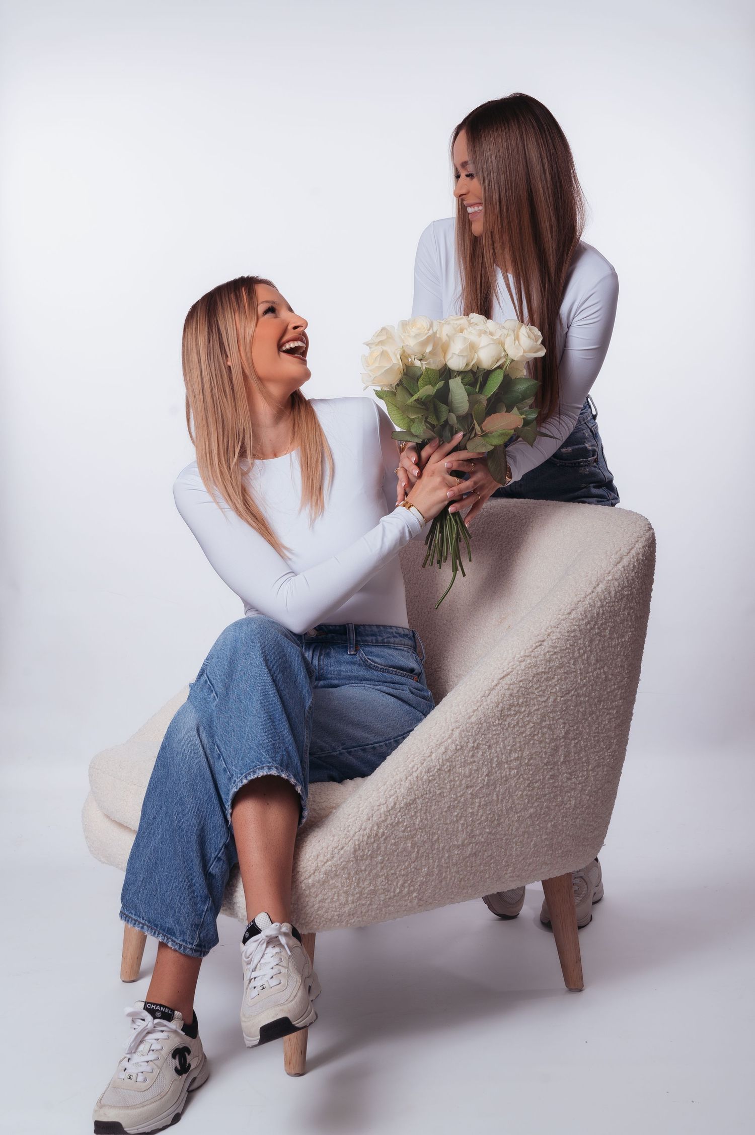 Two women in white tops and jeans share white roses, smiling joyfully in a bright studio setting.