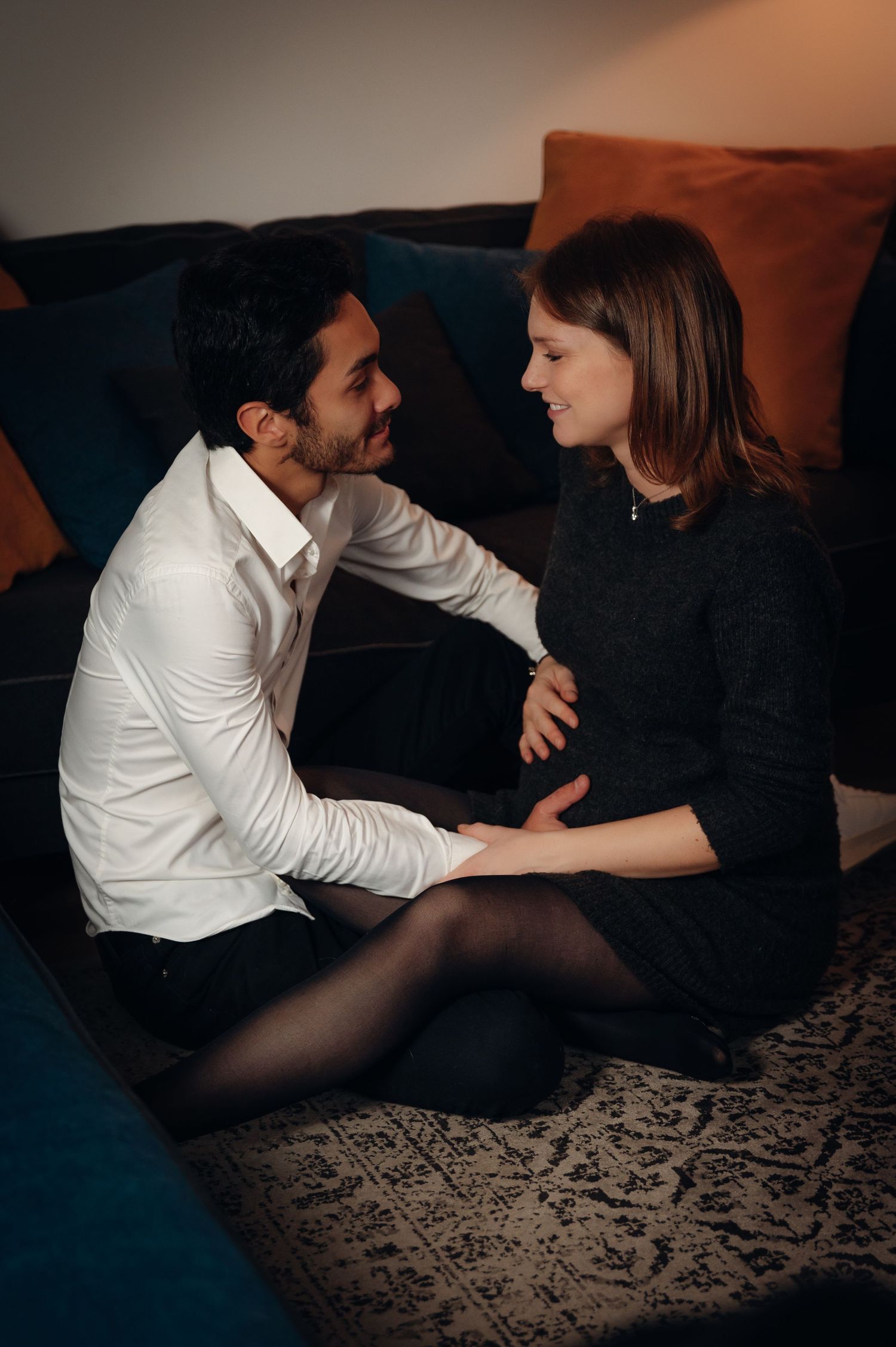 A couple sits on the floor leaning against a sofa, smiling at each other in a warmly lit living room.