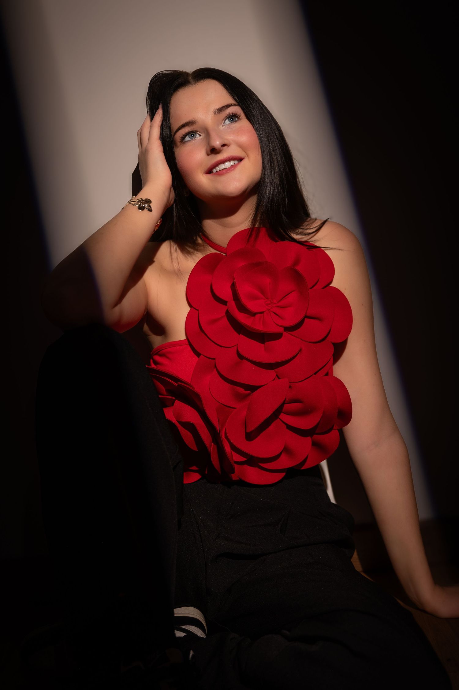 Young woman in red floral appliqué top and black pants poses elegantly against dramatic dark background.