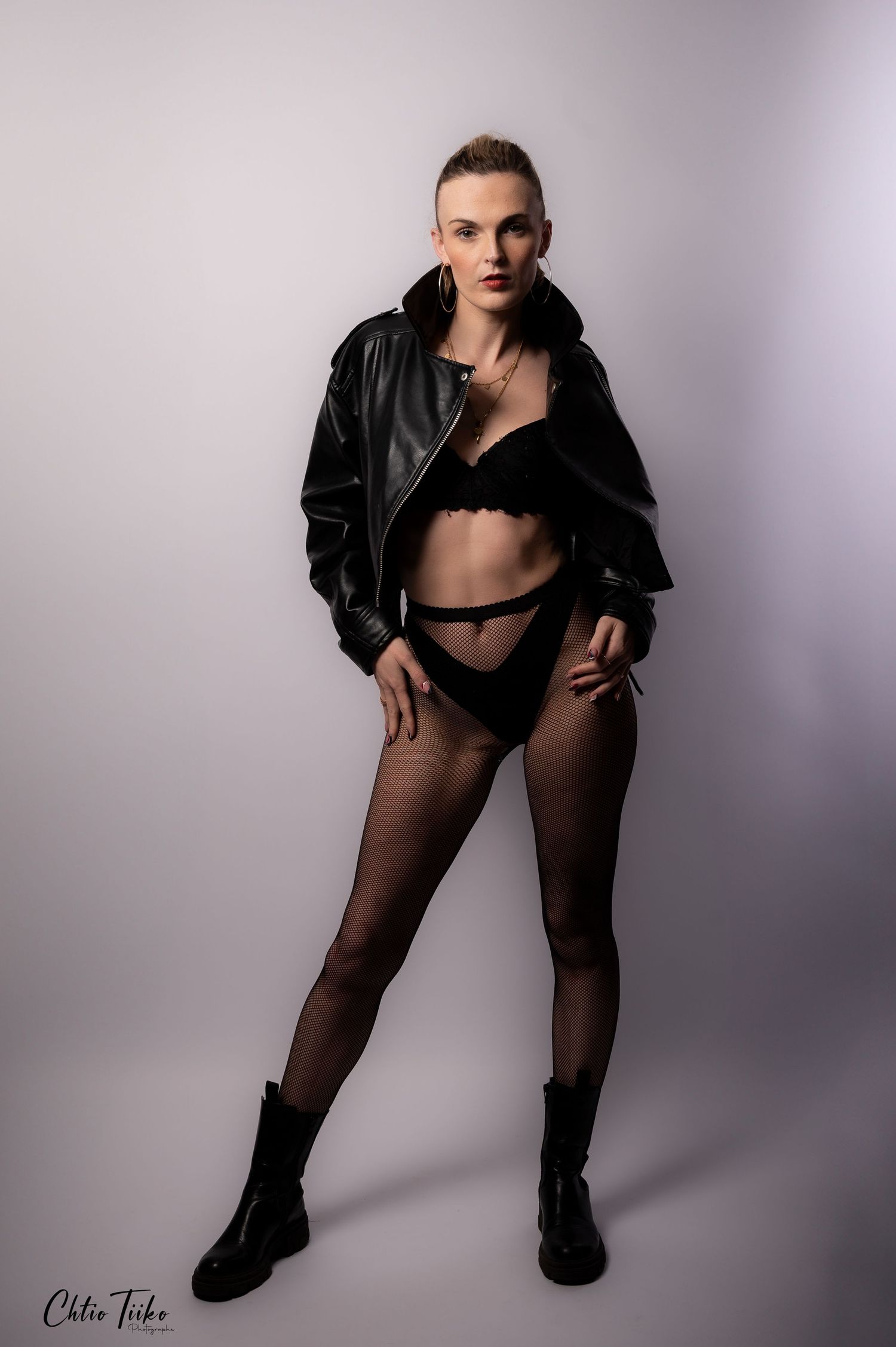 Woman in black lingerie, leather jacket, fishnet tights, and combat boots posing confidently in a studio.